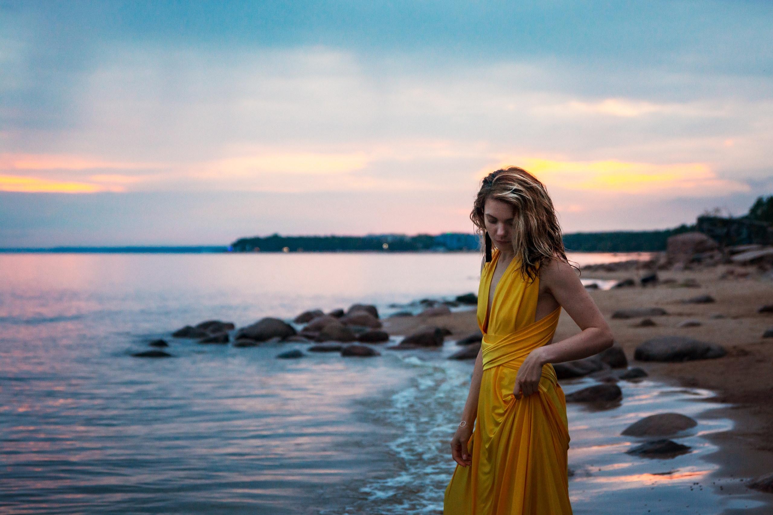 Фотография запечатлевает момент из фотосессии, где девушка в желтом платье на фоне заката наслаждается процессом позирования на камнях берега Финского залива.
