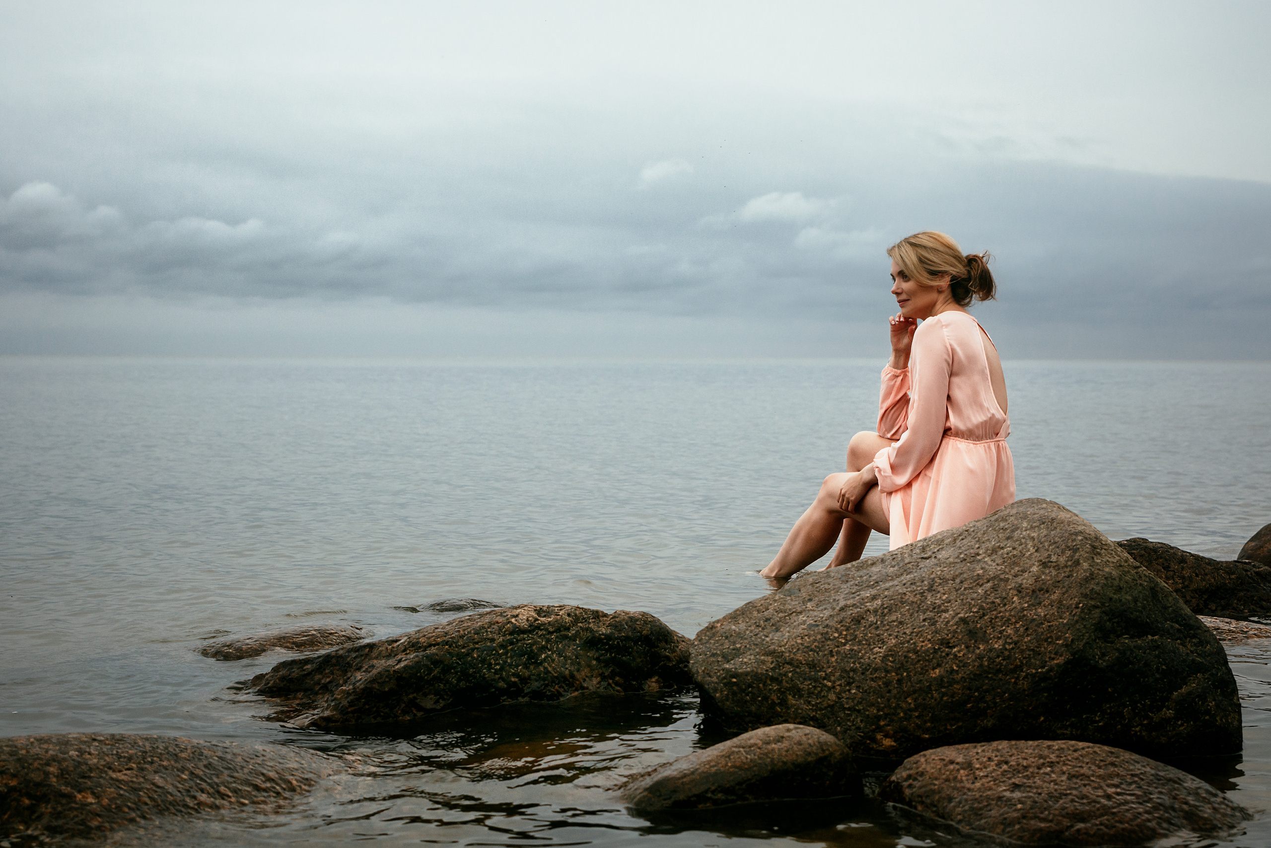 Красота водной глади отражается вокруг девушки, которая грациозно сидит на камнях в своем розовом платье.