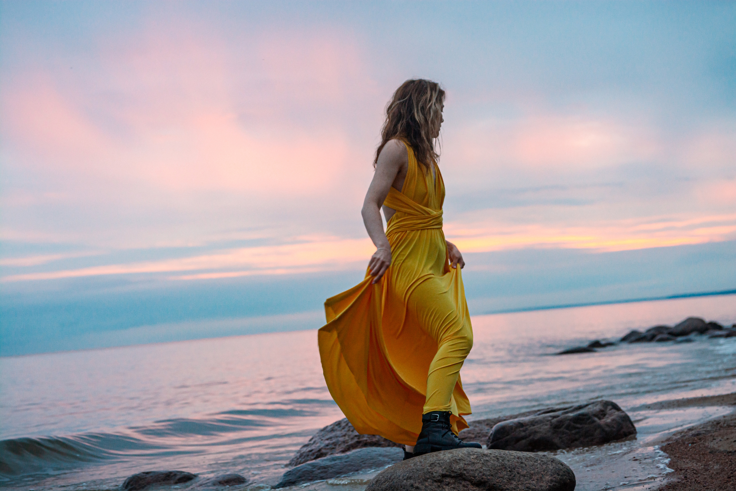 Силуэт девушки в желтом платье окутывает закатные лучи, пока она легко пышет на ветру на камнях берега Финского залива.