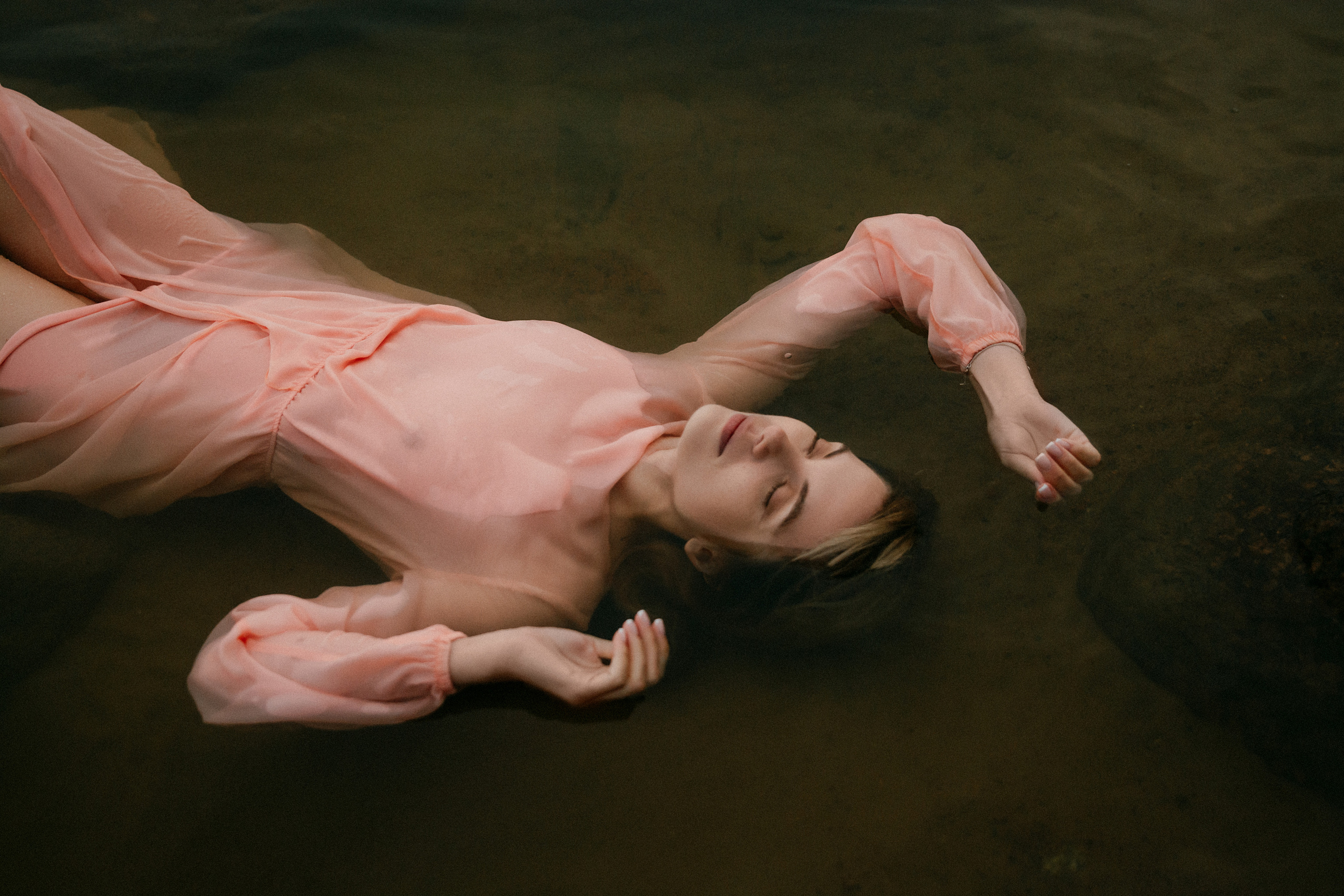 Девушка в легком розовом платье лежит в воде, воплощая умиротворение и покой.