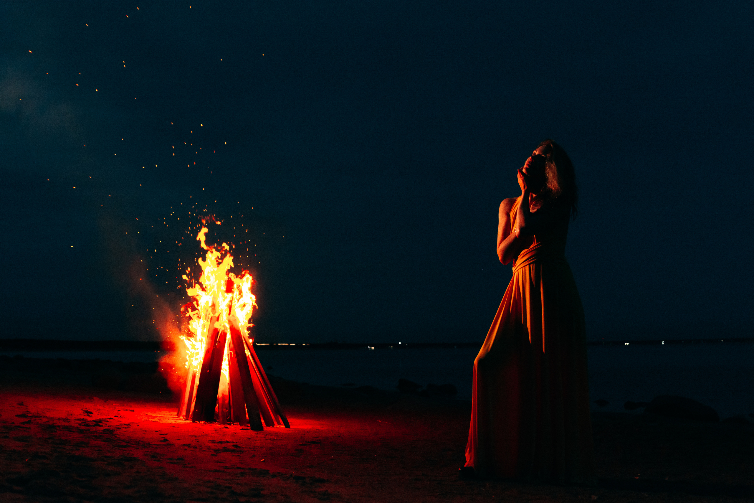 Костер на заднем плане создает эффектный контраст с девушкой, которая стоит в желтом платье, подчеркивая ее красоту и грацию.