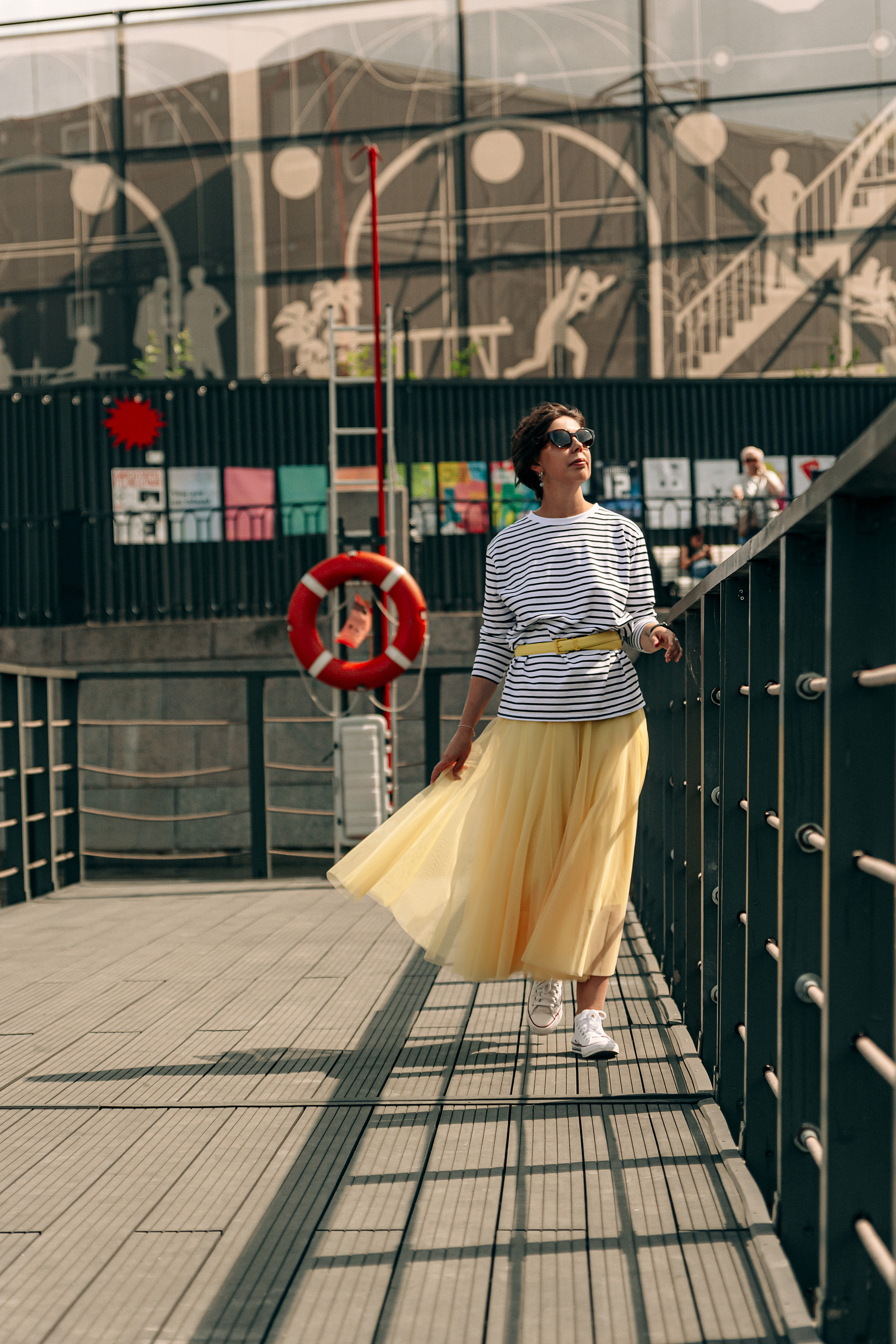 В живописной окружении Петербурга, девушка в жёлтой юбке и тельняшке создаёт яркую и модную картину, гуляя по Новой Голландии.