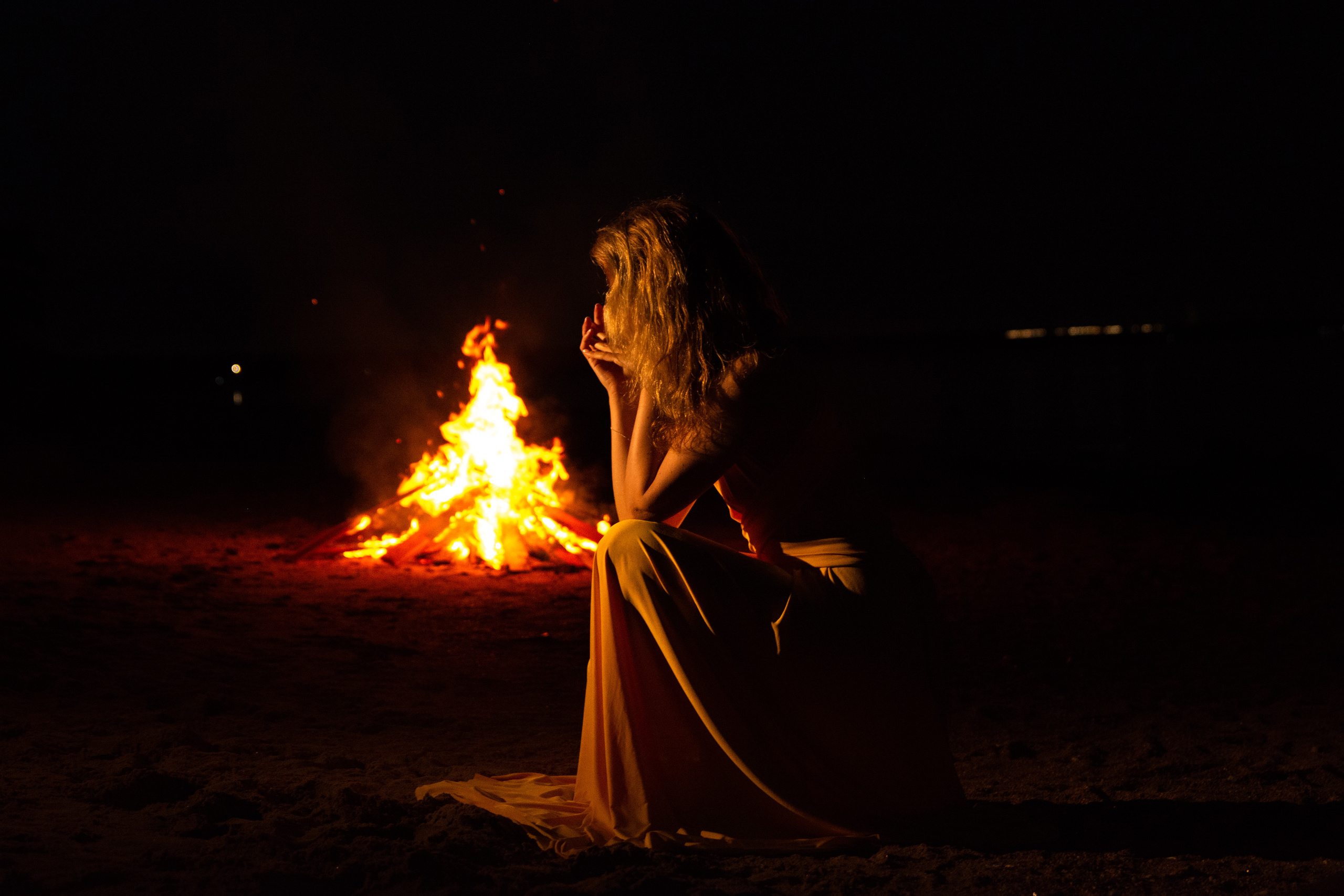 Силуэт девушки в желтом платье выделяется на фоне теплого огня костра, создавая загадочную и атмосферную композицию.