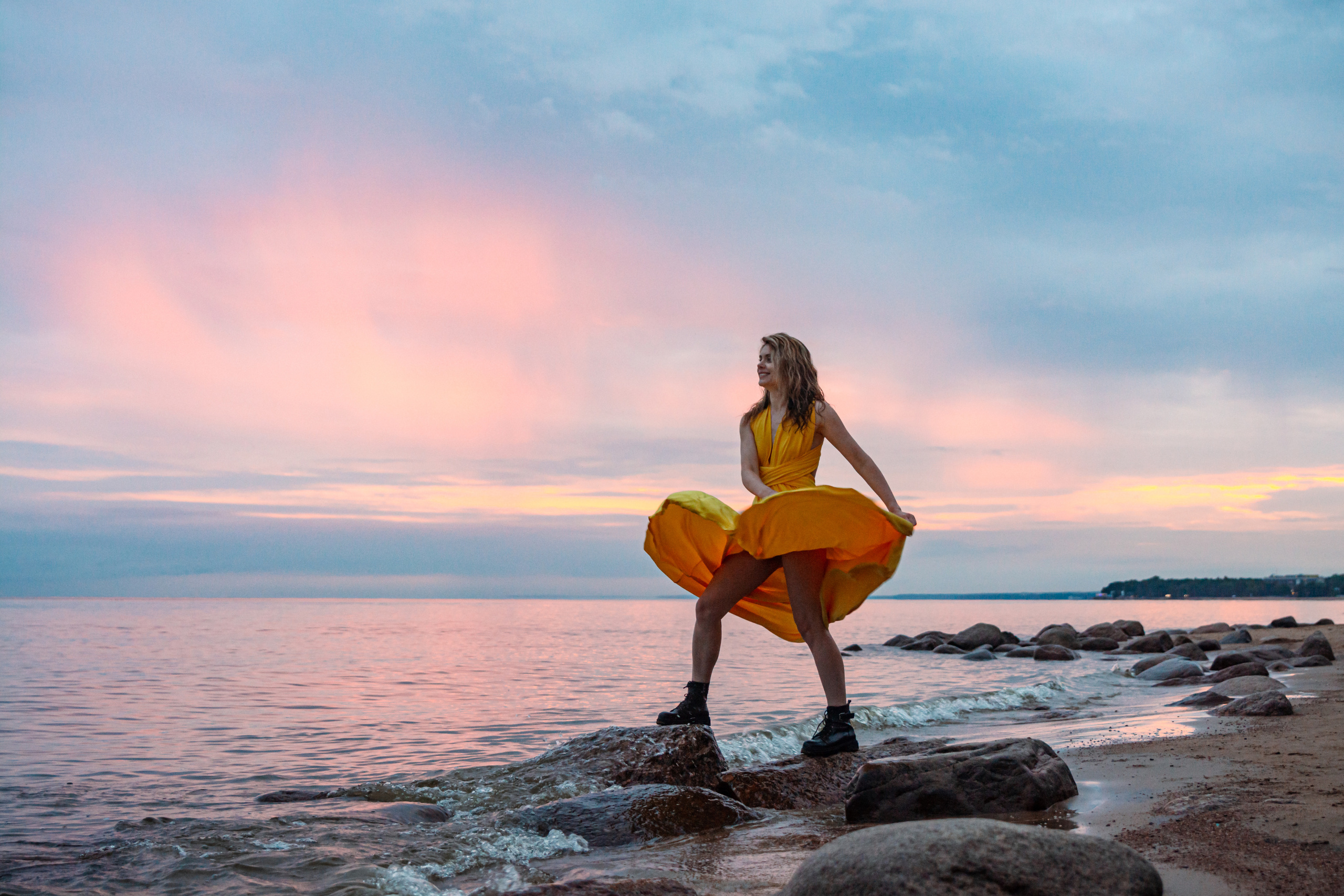 Девушка в желтом платье танцует на камнях берега Финского залива перед магическим закатом.