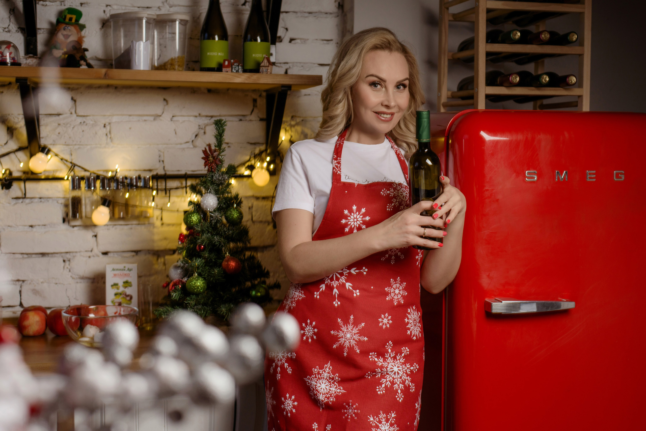 Фотосессия девушки блондинки в красном фартуке на кухне около красного холодильника с бутылкой вина на новый год