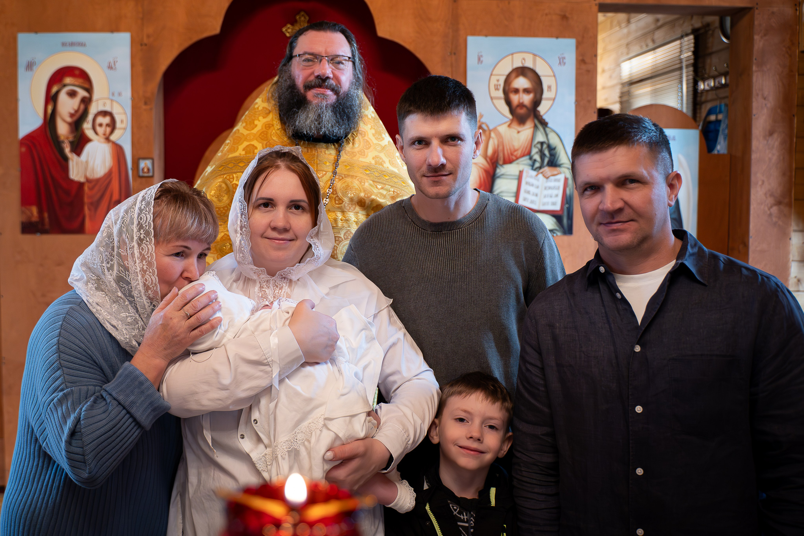 Счастливые родители с ребенком на руках и крестные стоят у алтаря после завершения обряда крещения.   Семья позирует фотографу на крыльце деревянной церкви после обряда крещения