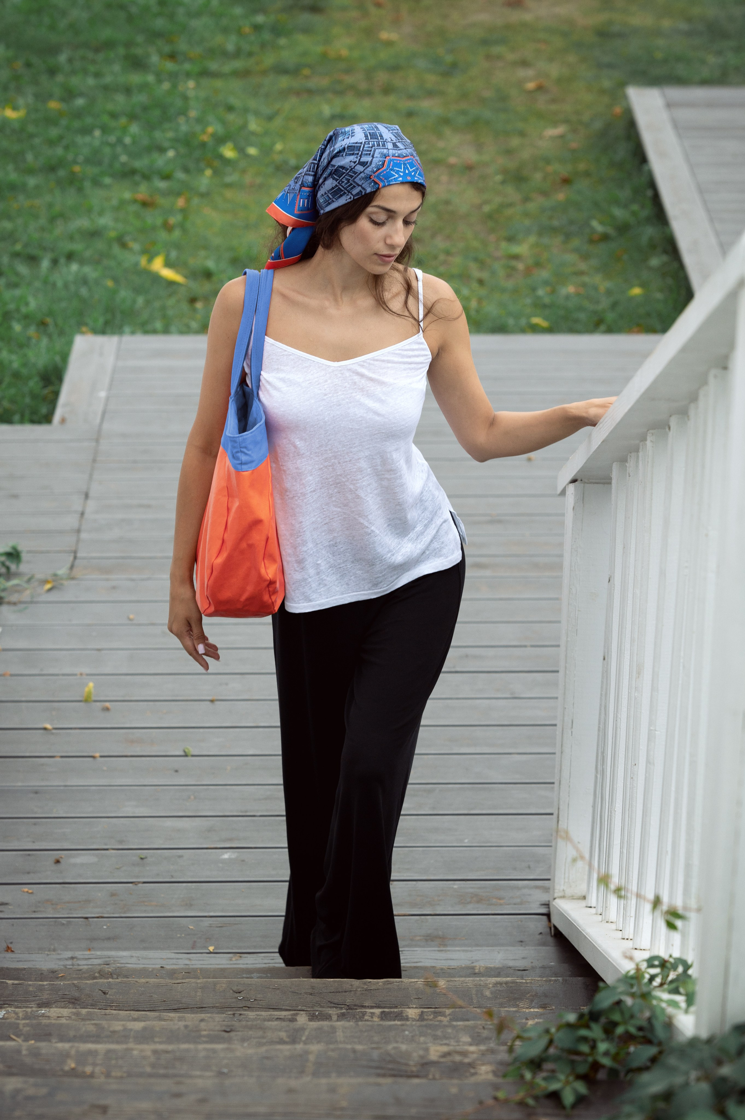 Красивая брюнетка в белом топе и черных брюках поднимается по лестнице. На плече у нее оранжевая пляжная сумка, а на голове яркий синий платок.