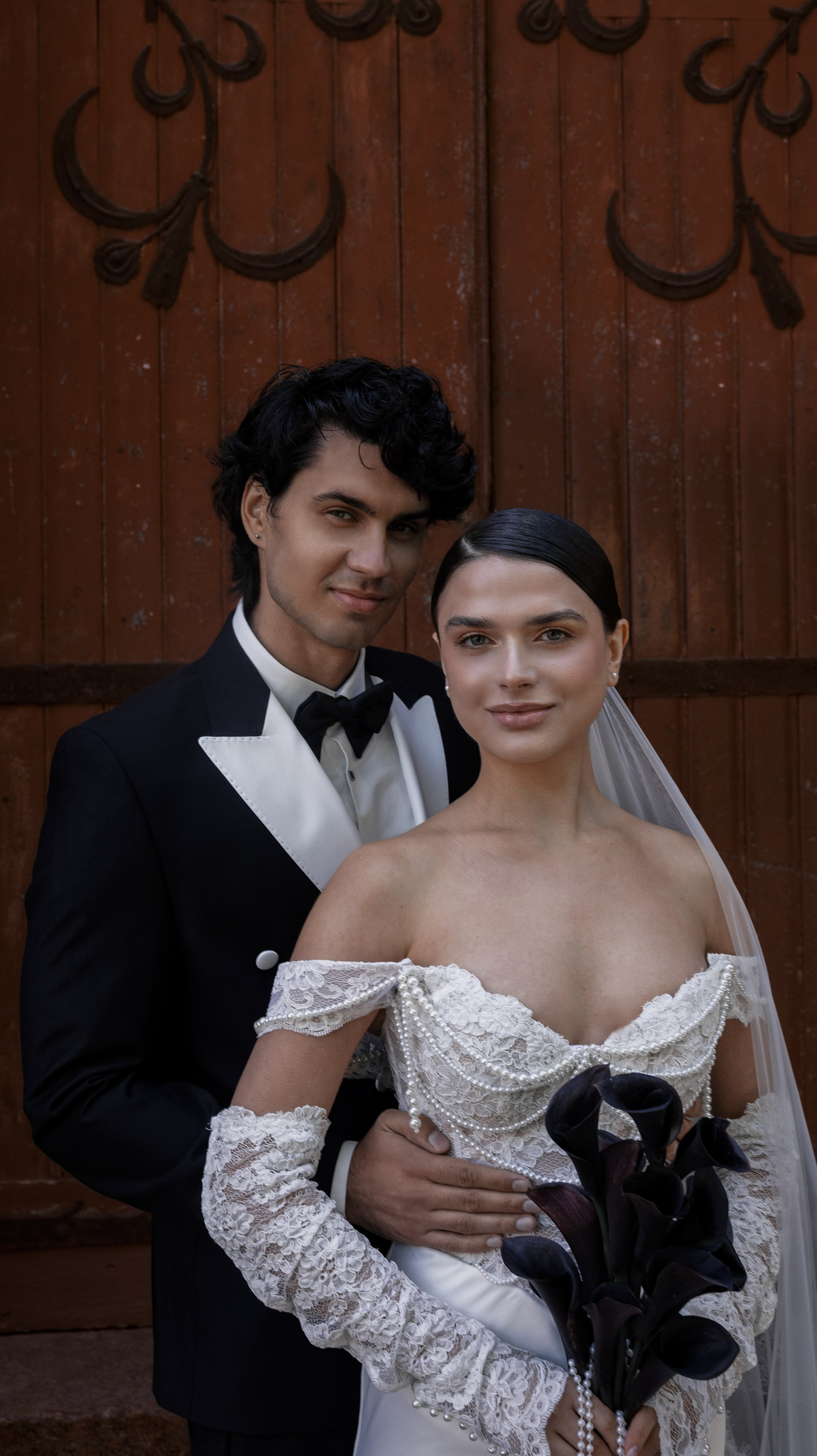 «Элегантный свадебный портрет невесты в платье с открытыми плечами, расшитом жемчугом, и кружевных перчатках, держащей в руках черные каллы, стоящей рядом с женихом в черном смокинге с белыми лацканами на фоне деревенской деревянной двери».