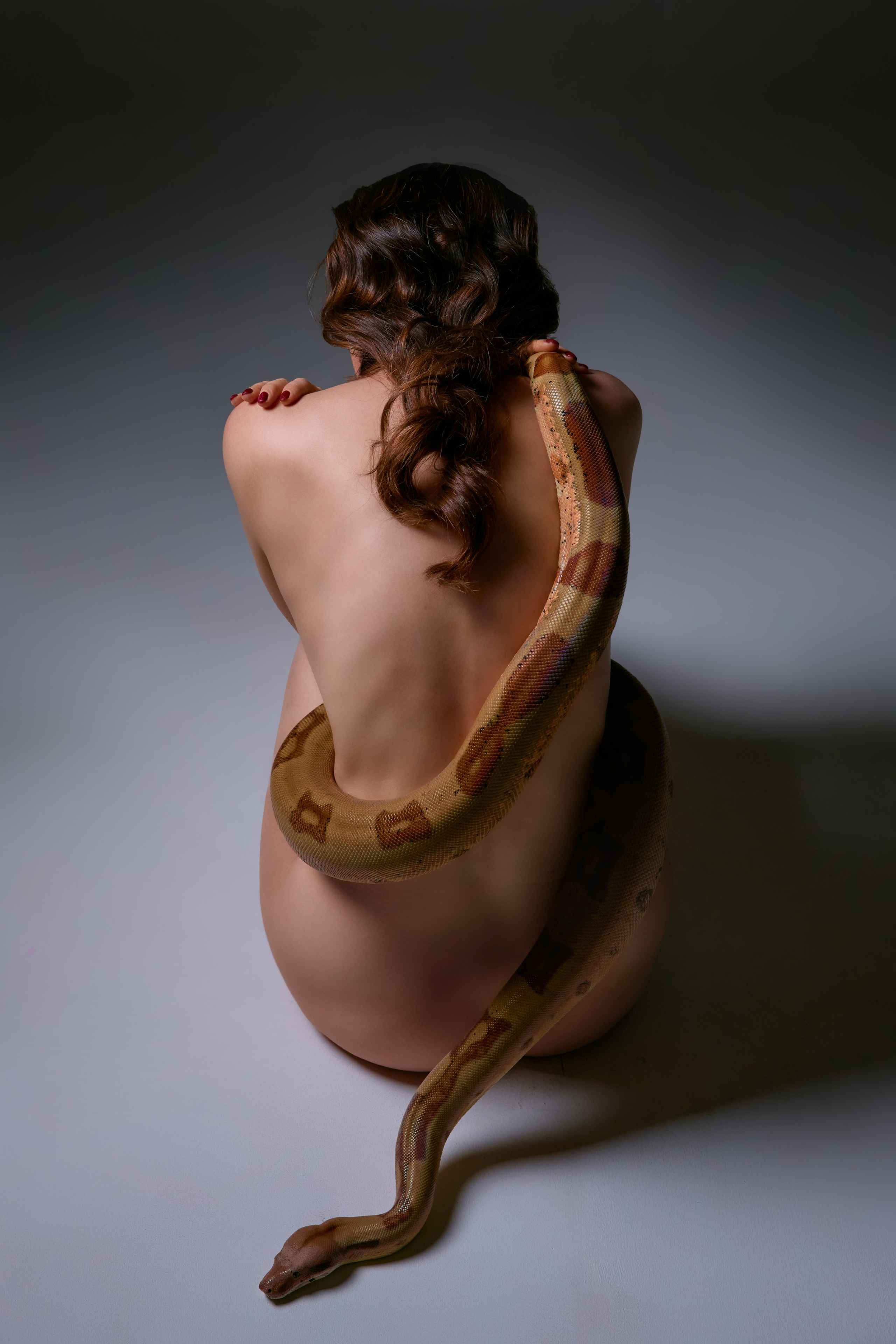 На фотографии изображена художественная композиция, объединяющая естественную красоту человеческого тела и дикую эстетику природы. Обнажённая женская фигура, освещённая мягким рассеянным светом, гармонично переплетается с извивающейся змеёй, чья гладкая чешуя контрастирует с нежными линиями кожи. Цветовая гамма выдержана в приглушённых тонах — золотистых, терракотовых и глубоких зелёных оттенках, что подчёркивает вневременной и символический характер изображения. Поза модели и движение змеи создают динамику, отражая тему единства противоположностей: хрупкости и силы, свободы и опасности. Фотография передаёт атмосферу загадочности и первобытной гармонии, акцентируя внимание на текстурах и плавных формах.