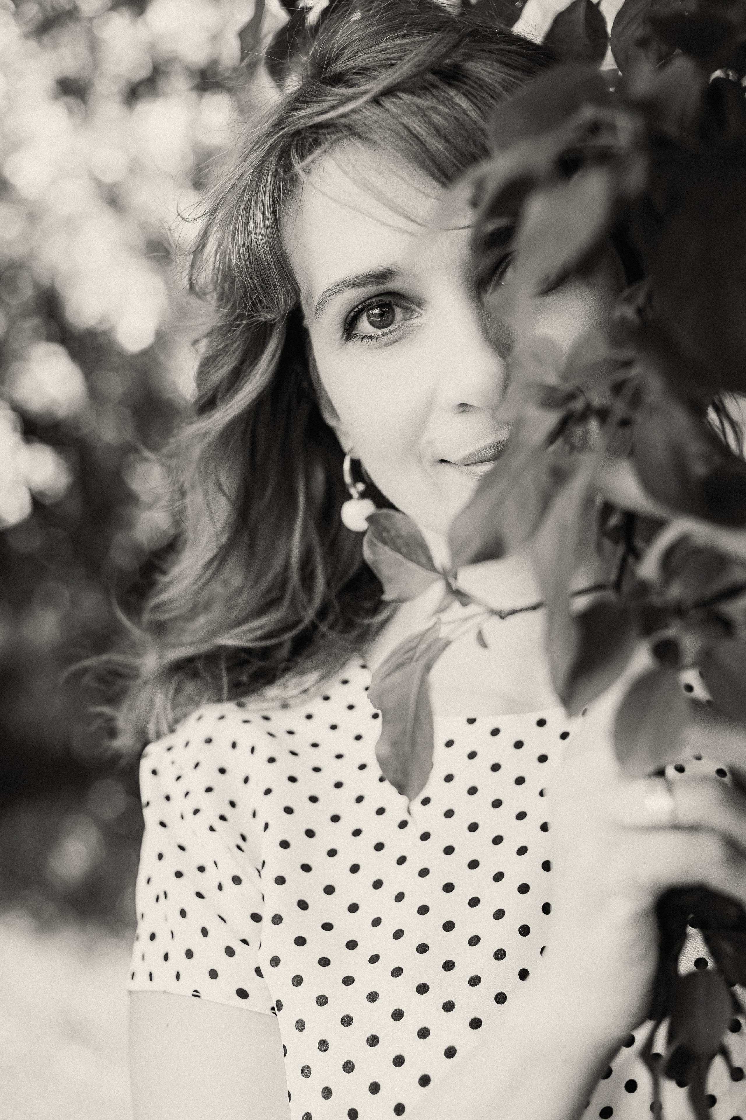 Черно-белый портрет молодой женщины в гороховом платье, которая стоит за ветвями с листьями и смотрит в камеру с легкой улыбкой.