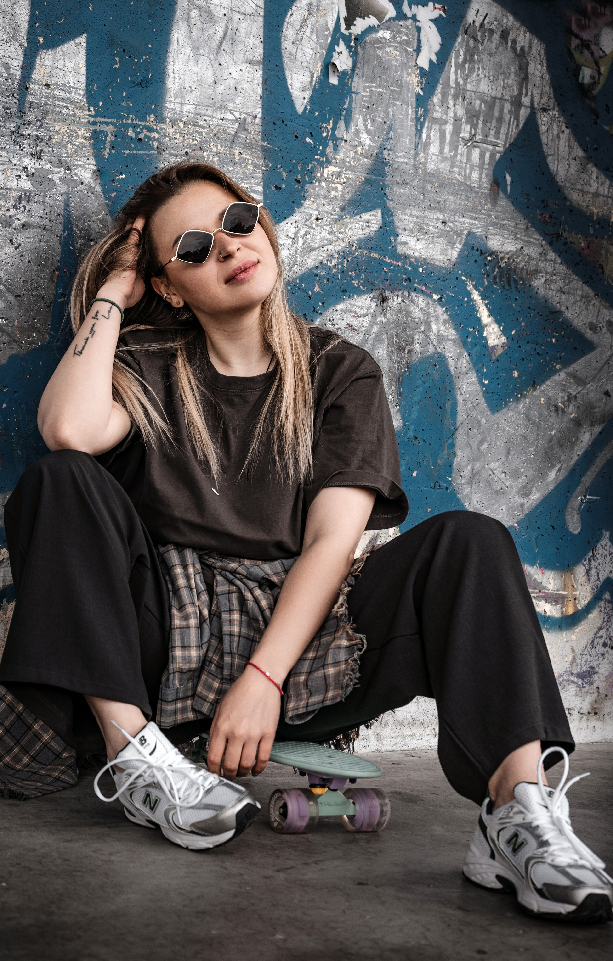 Эджи streetwear look — девушка с татуировкой и пирсингом сидит на мятном круизере, oversize чёрная футболка, широкие брюки, белые массивные New Balance, граффити-стена, расслабленная поза, скейт-культура.