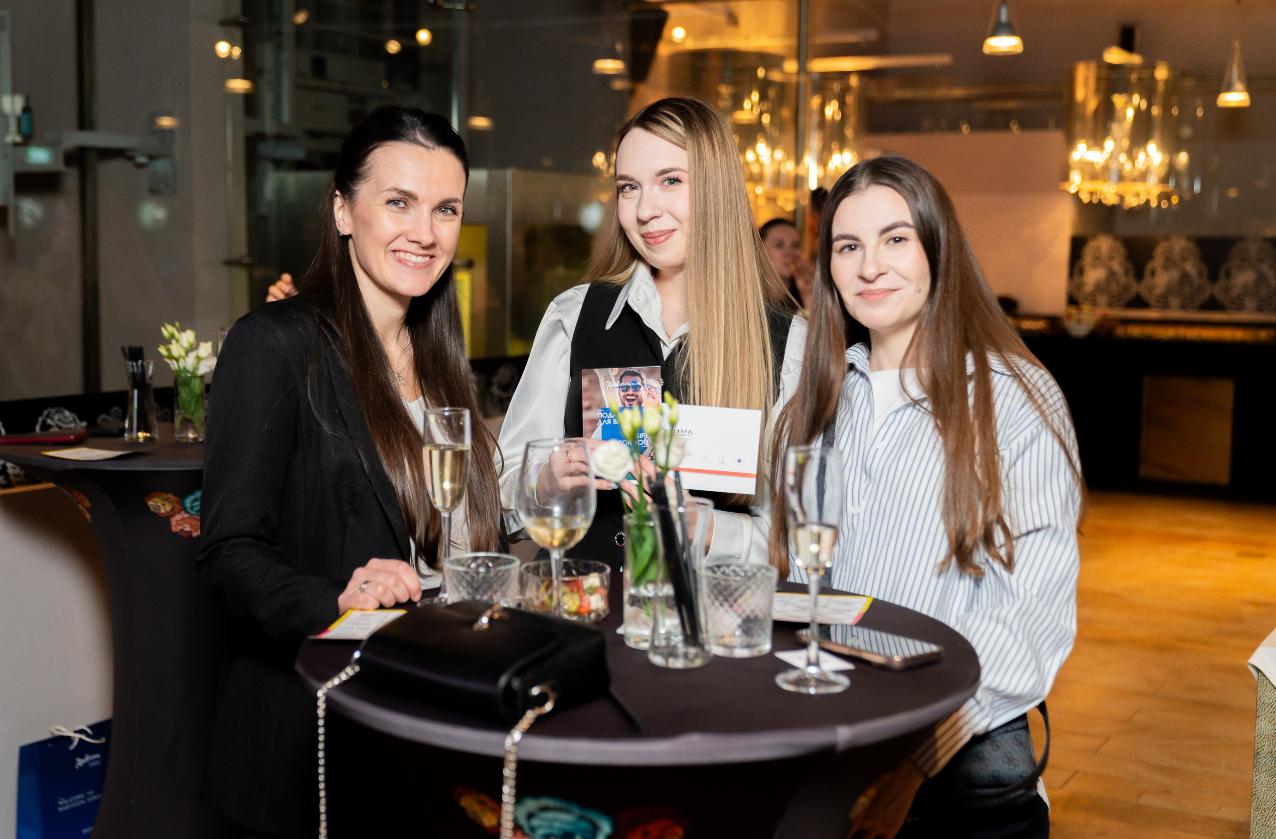 Три девушки с бокалами шампанского и подарочным сертификатом за столом на вечере в Radisson Sonya. Фотограф Евгений Мальцев.
