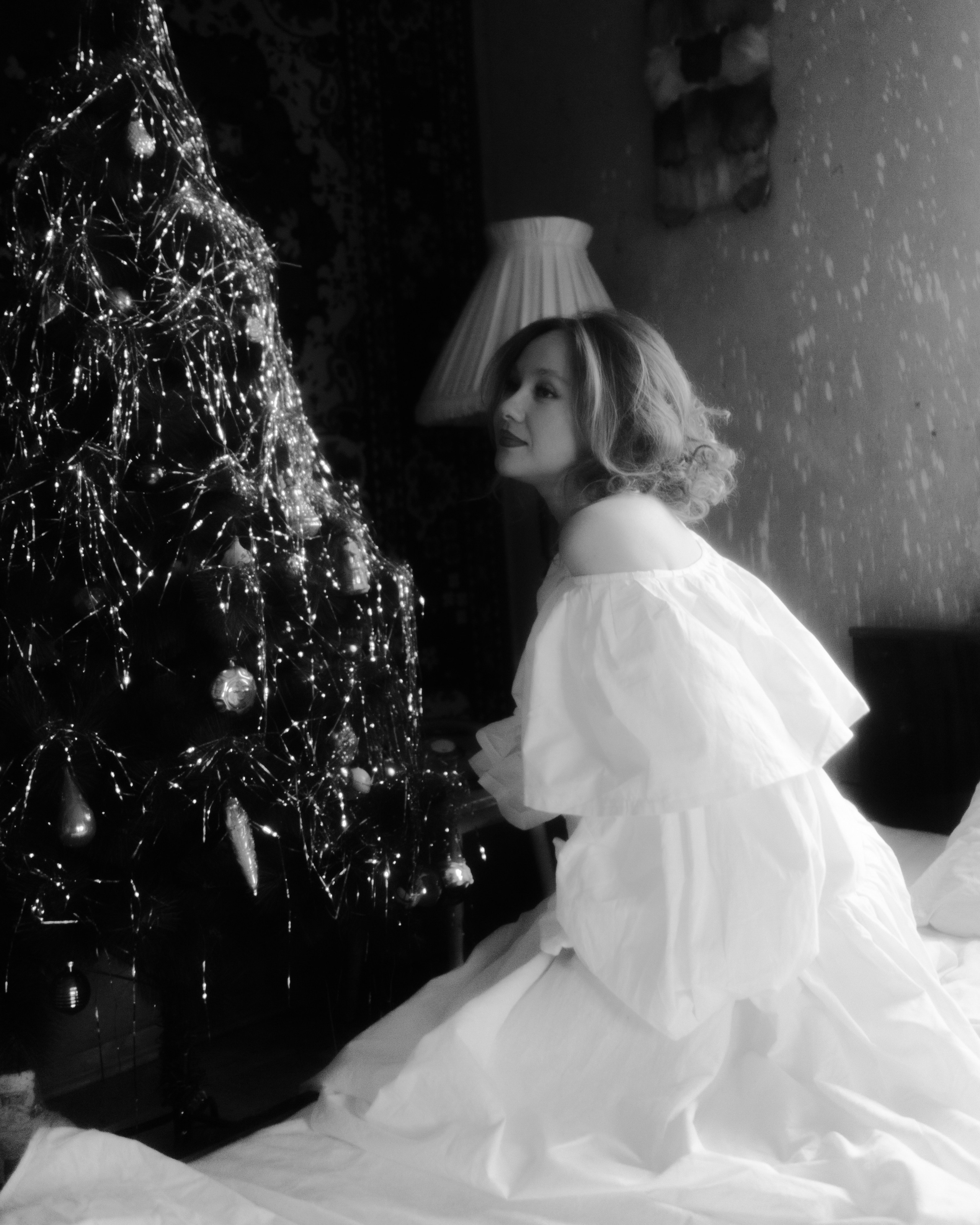 Чб портрет у елки: беременная в белом платье на кровати, винтажная лампа — атмосферная новогодняя фотосессия беременности в Санкт-Петербурге.