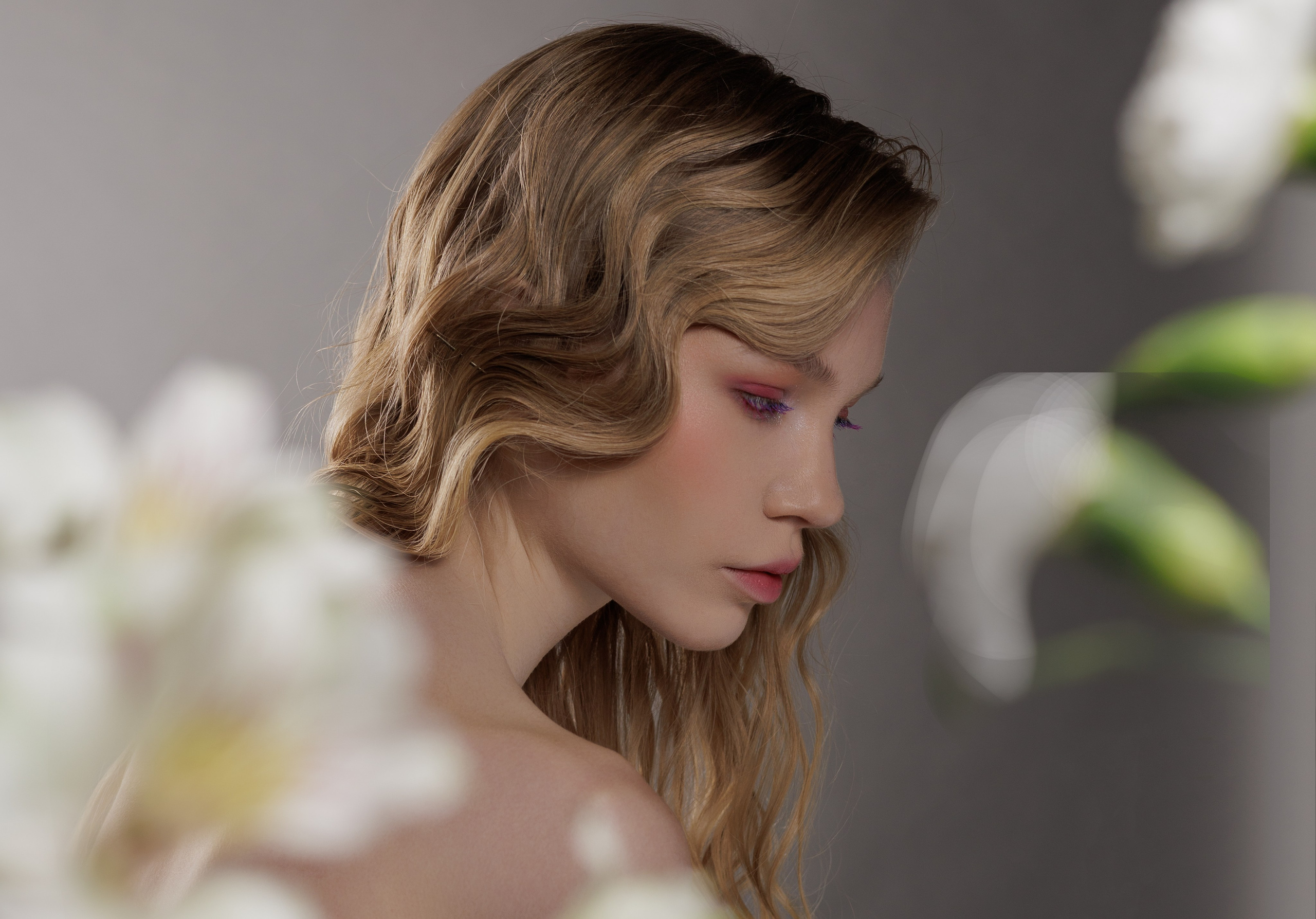 Женщина с волнистыми светлыми волосами задумчиво смотрит вниз. Цветы с мягким акцентом обрамляют ее, создавая безмятежное, созерцательное настроение на нейтральном фоне.