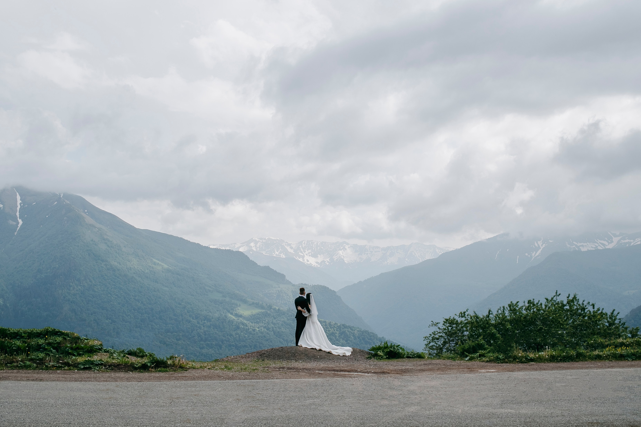 Свадебный фотосет в горах, когда жених и невеста идут по тропинке, окруженной зелеными деревьями и цветами