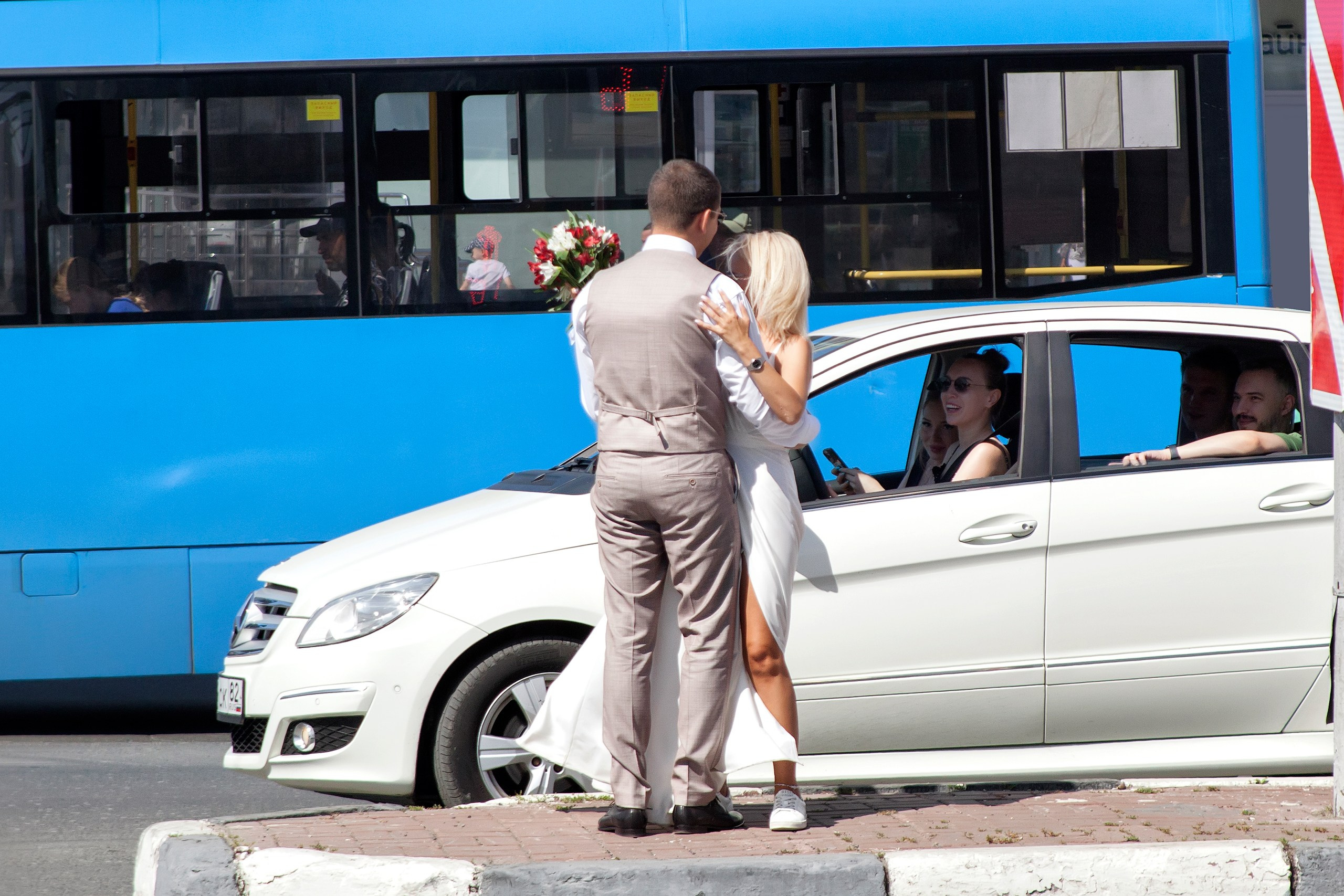 Жених и невеста на перекрёстке в городе на фоне проезжающего мимо синего автобуса, рядом остановилась машина и люди улыбаются молодым
