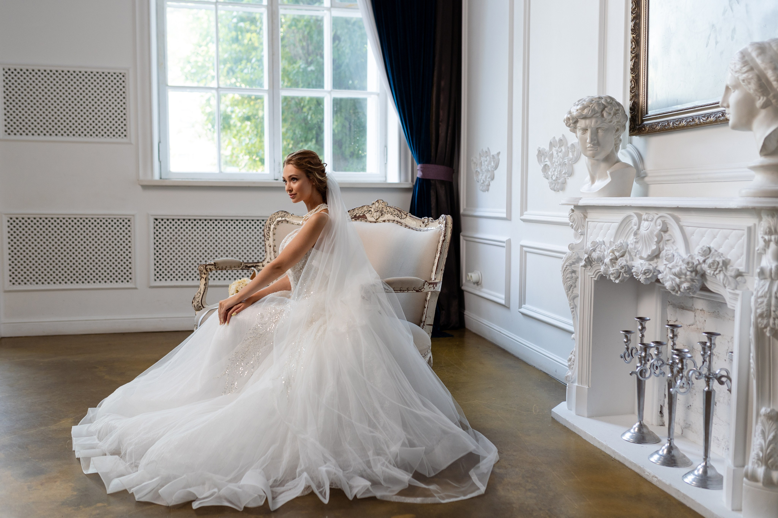 Невеста в длинном белом платье сидит на стуле на фоне окна.