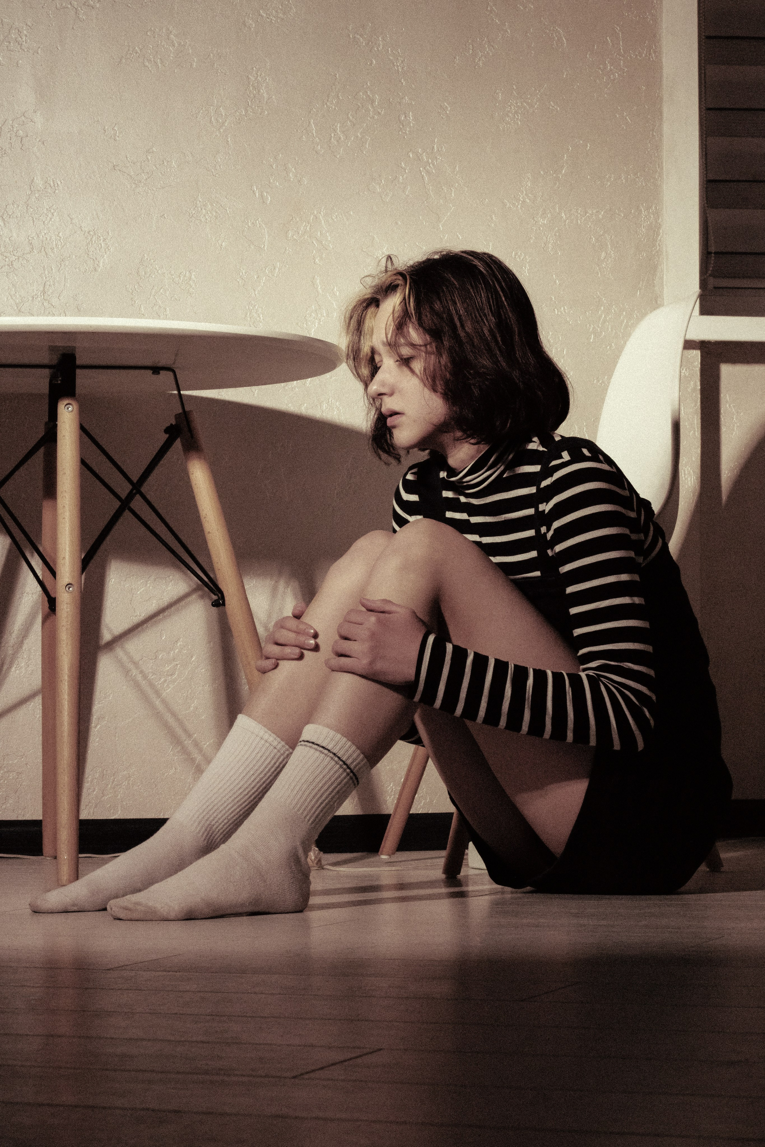 Девушка сидит на полу, обняв колени, рядом пустой стол — тишина становится невыносимой.