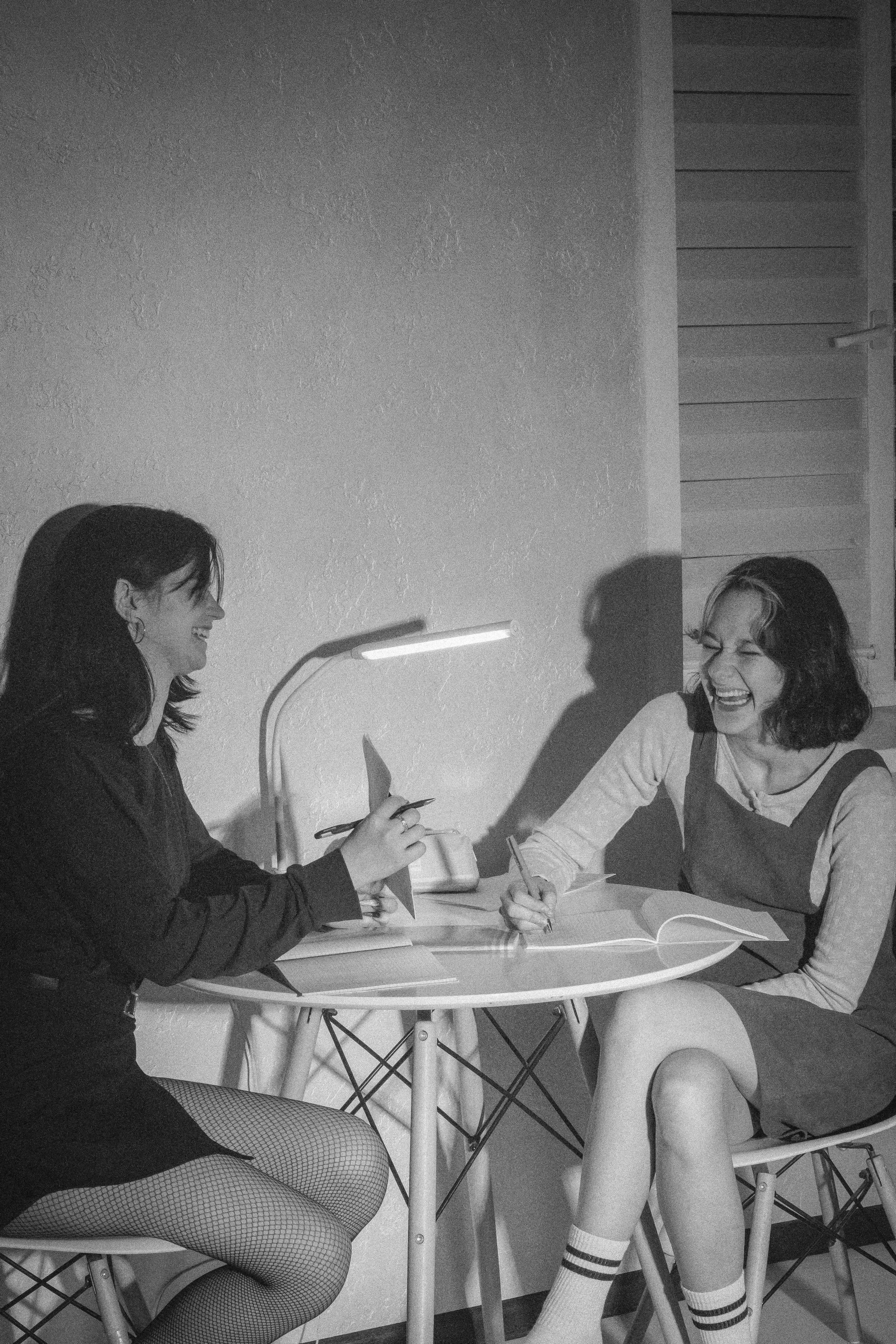 Две девушки смеются за столом, подсвеченные настольной лампой — момент, который теперь звучит эхом.