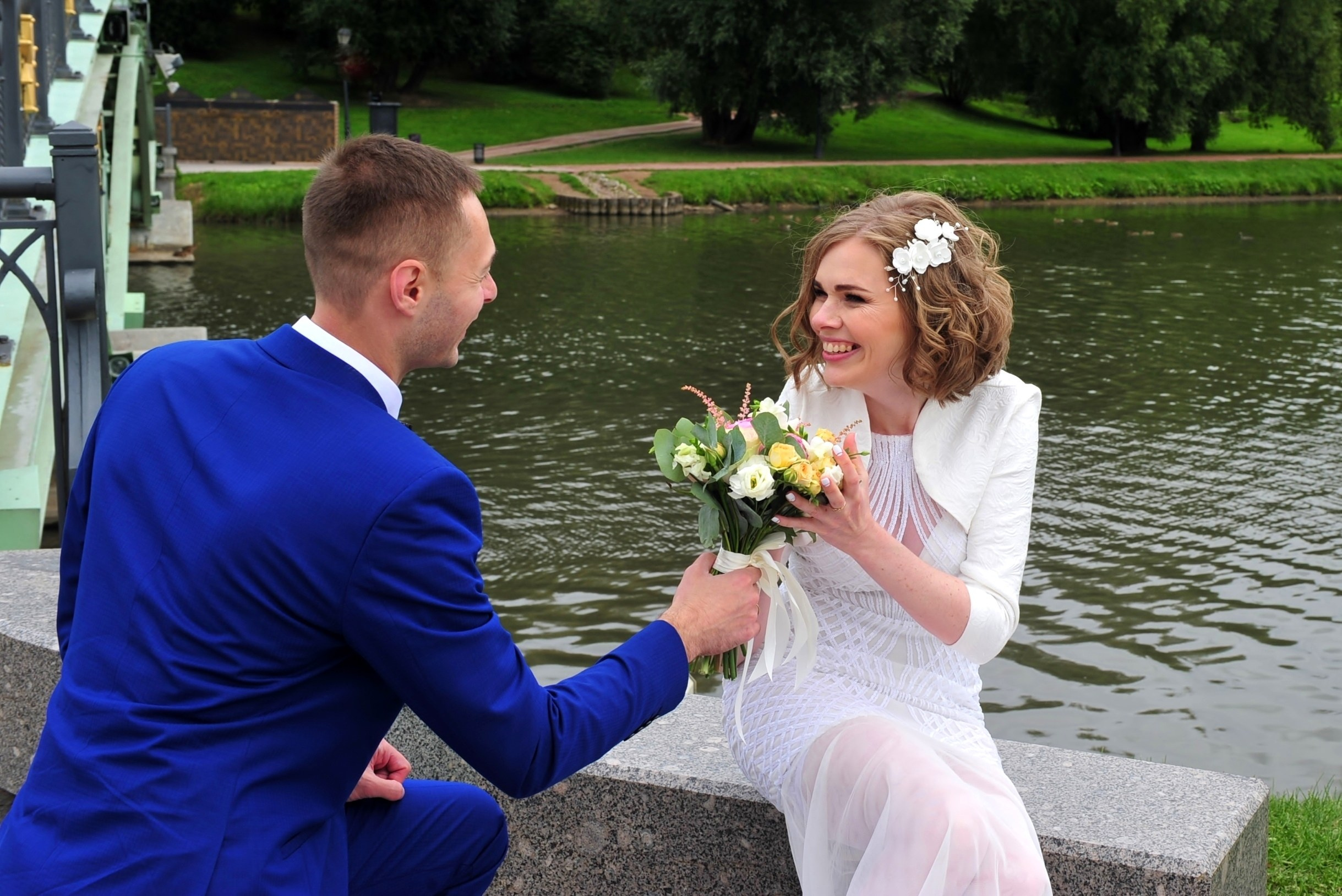 Свадьба в Царицыно хмурым прохладным летним днём, жених стоит на одном колене перед невестой и дарит ей букет, невеста счастлива и смеётся, молодожены на фоне пруда у моста