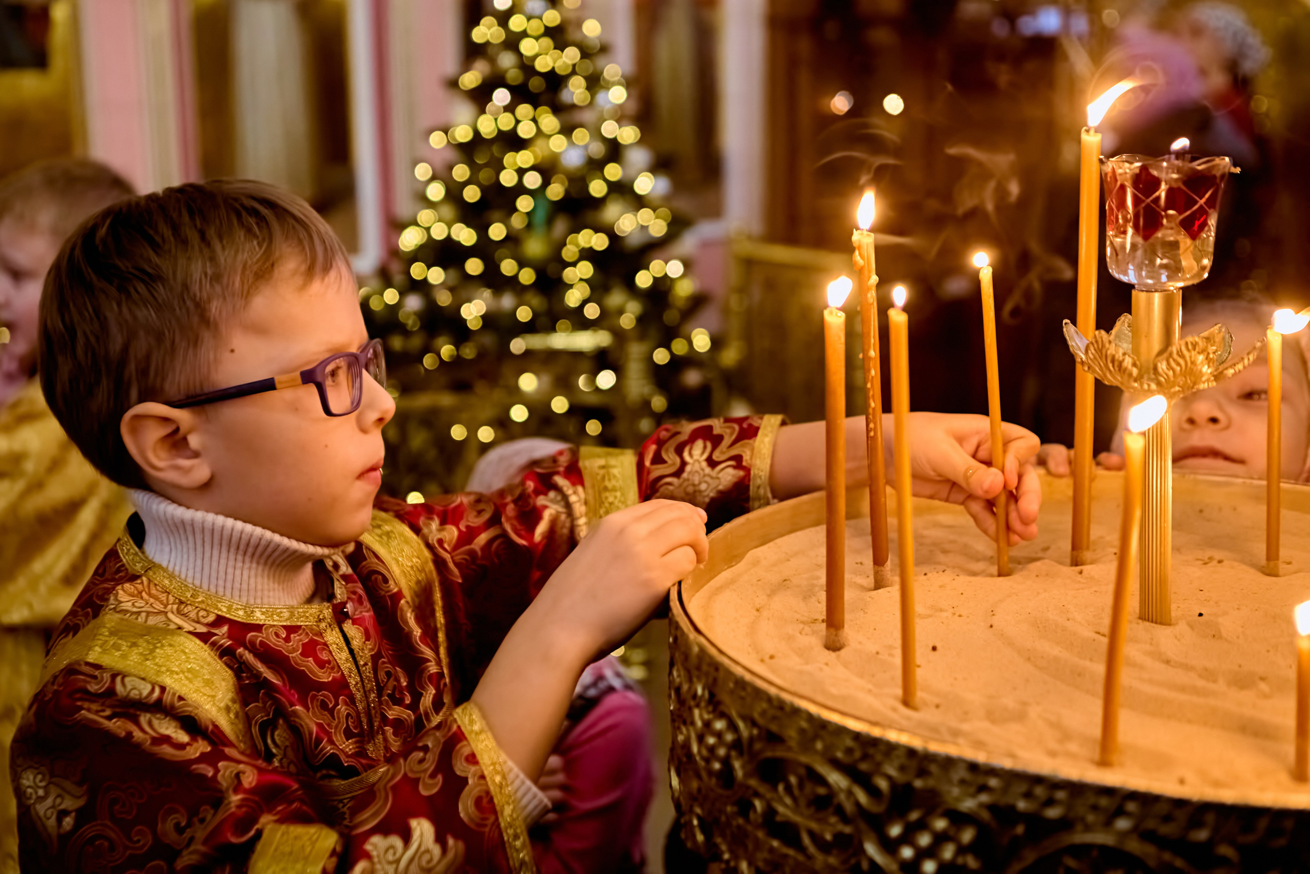 Маленький алтарник 8 лет в красном стихаре у подсвечника в храме на литургии