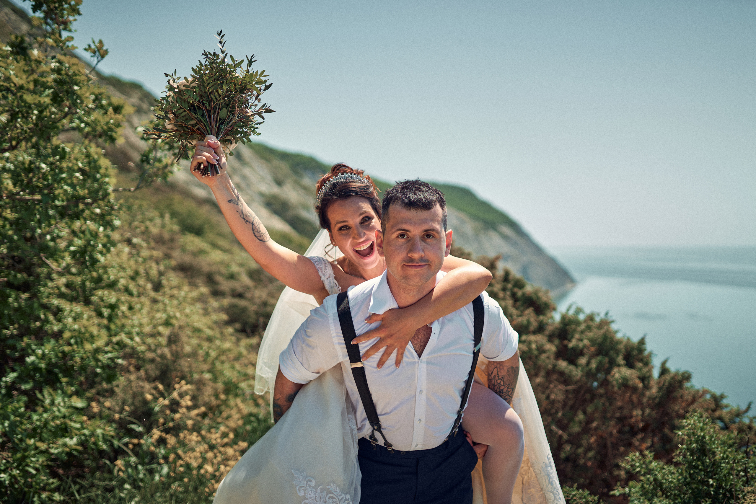 Невеста на спине у жениха улыбается и машет букетом