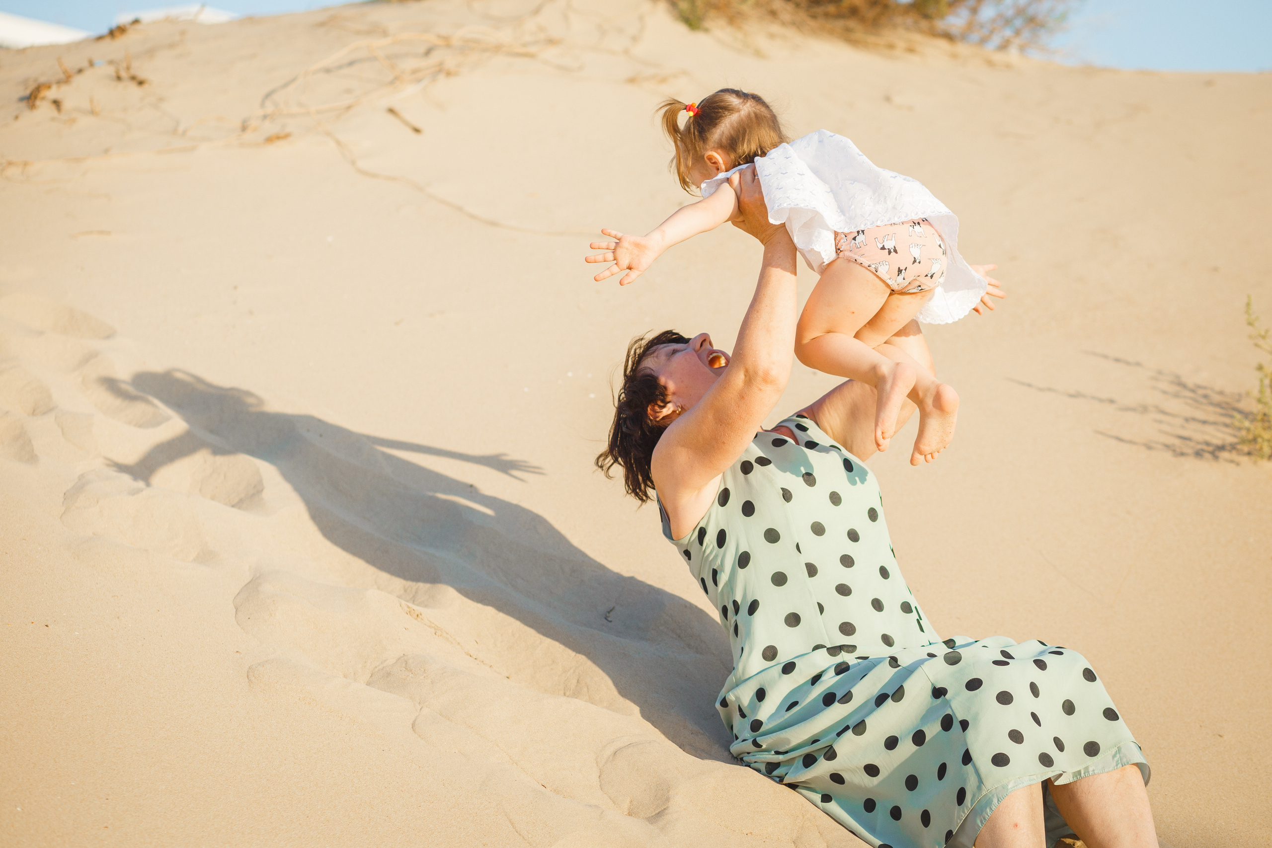 Бабушка поднимает внучку перед собой сидя на песке