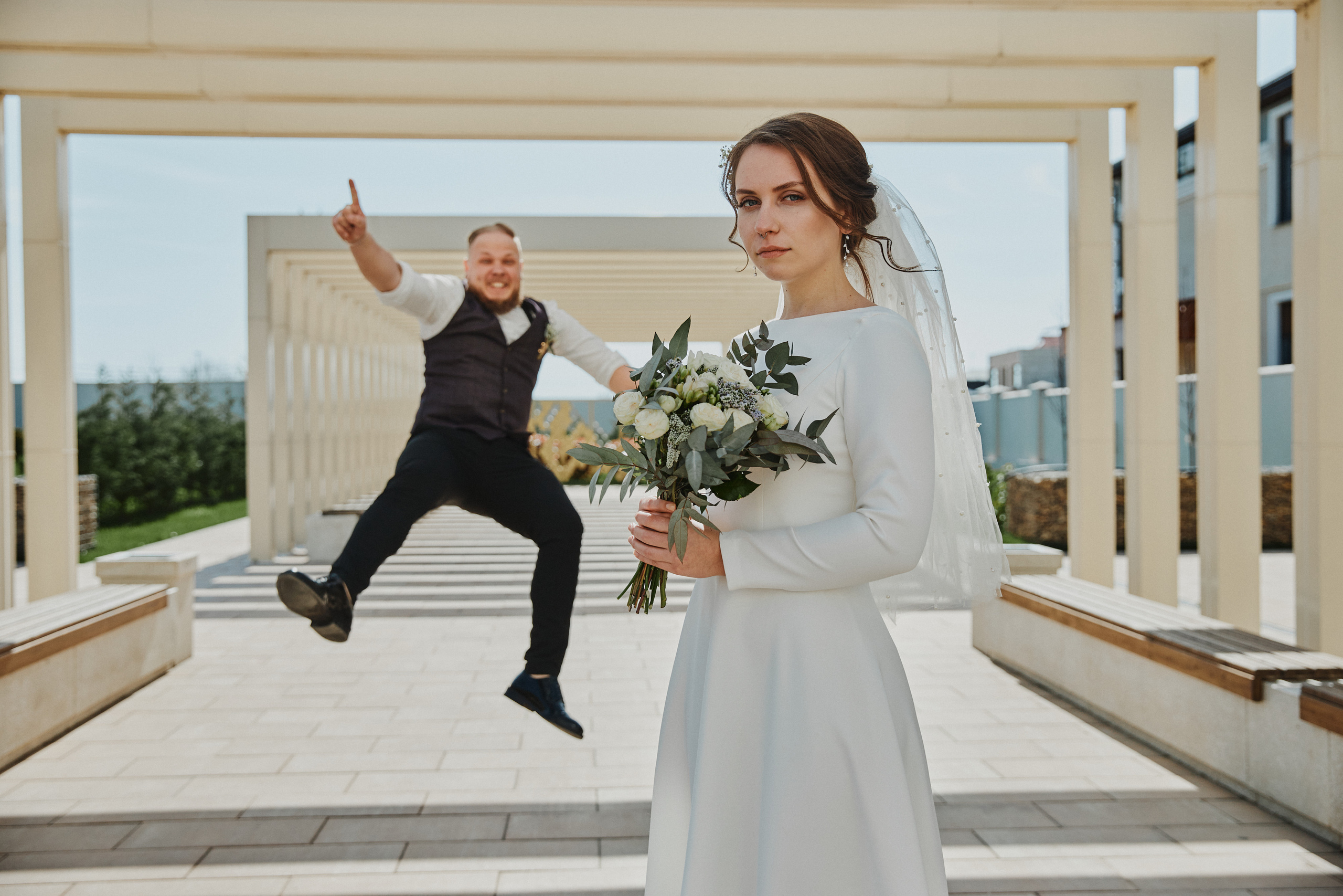 Жених прыгает на фоне невесты, пока они смотрит в камеру