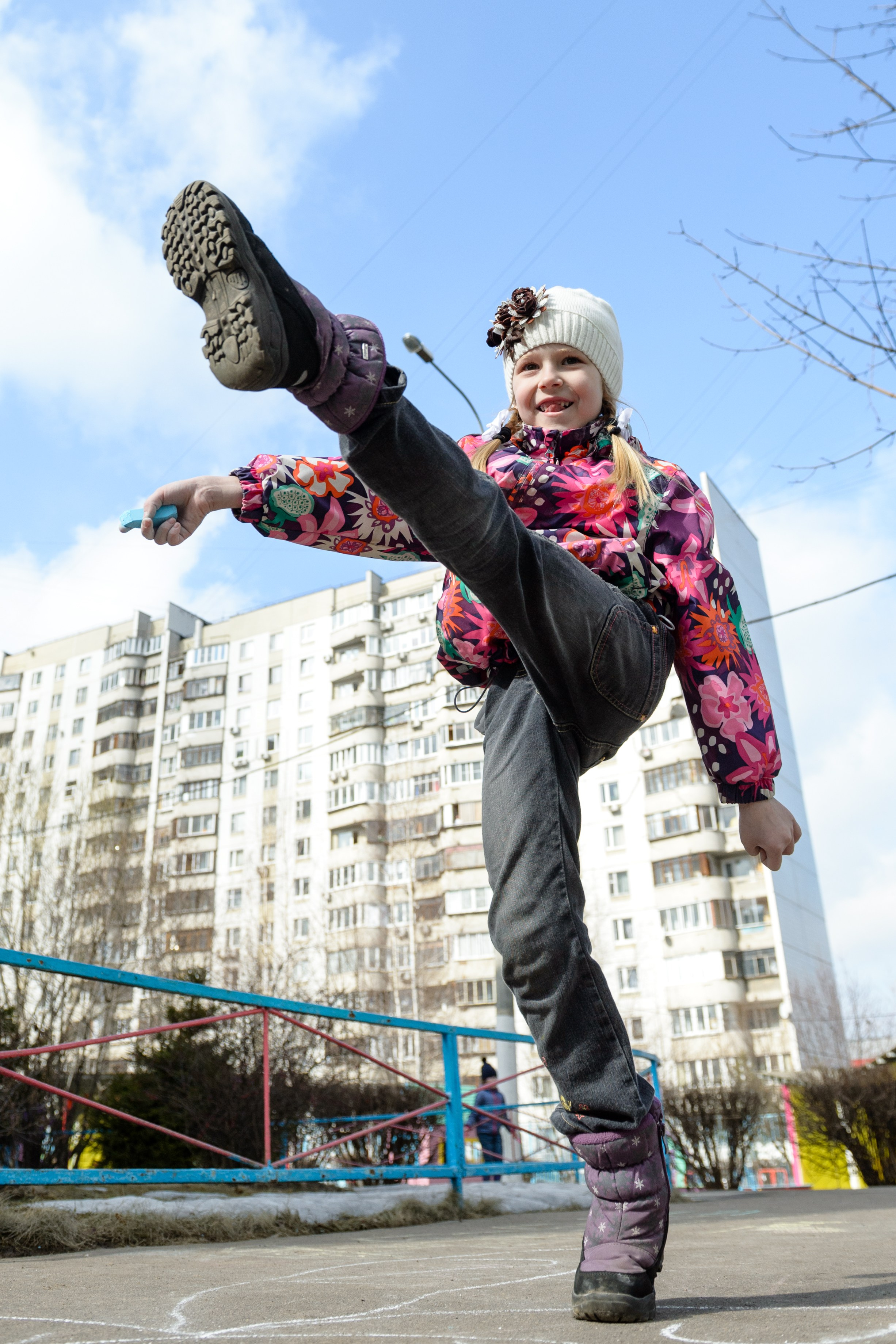 Девочка в зимней одежде делает высокий мах ногой на улице, динамичный кадр на фоне домов с нижнего ракурса.