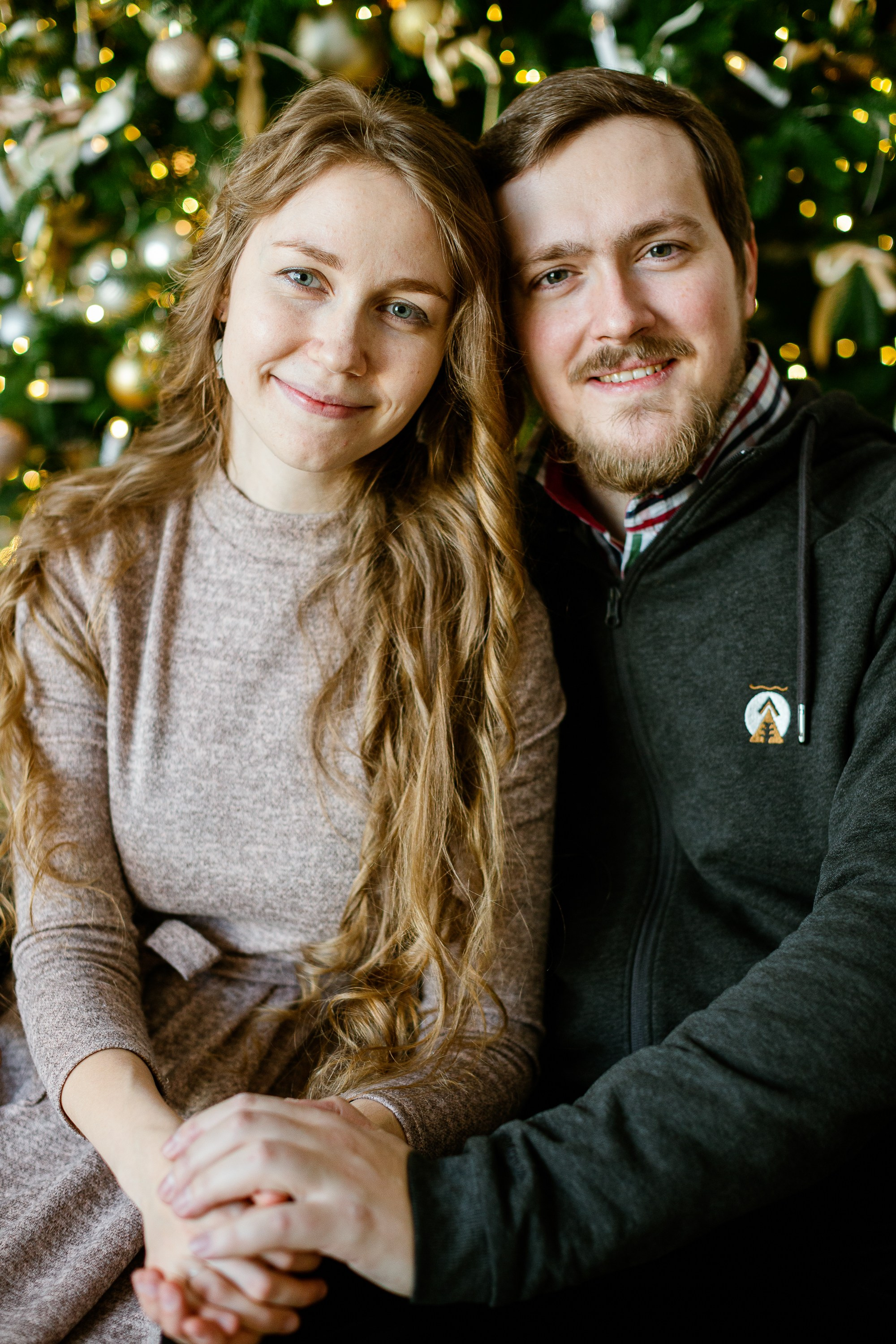 Семейная сидит на полу на фоне стоит елка и светится огоньками. Муж и жена смотрят в камеру.