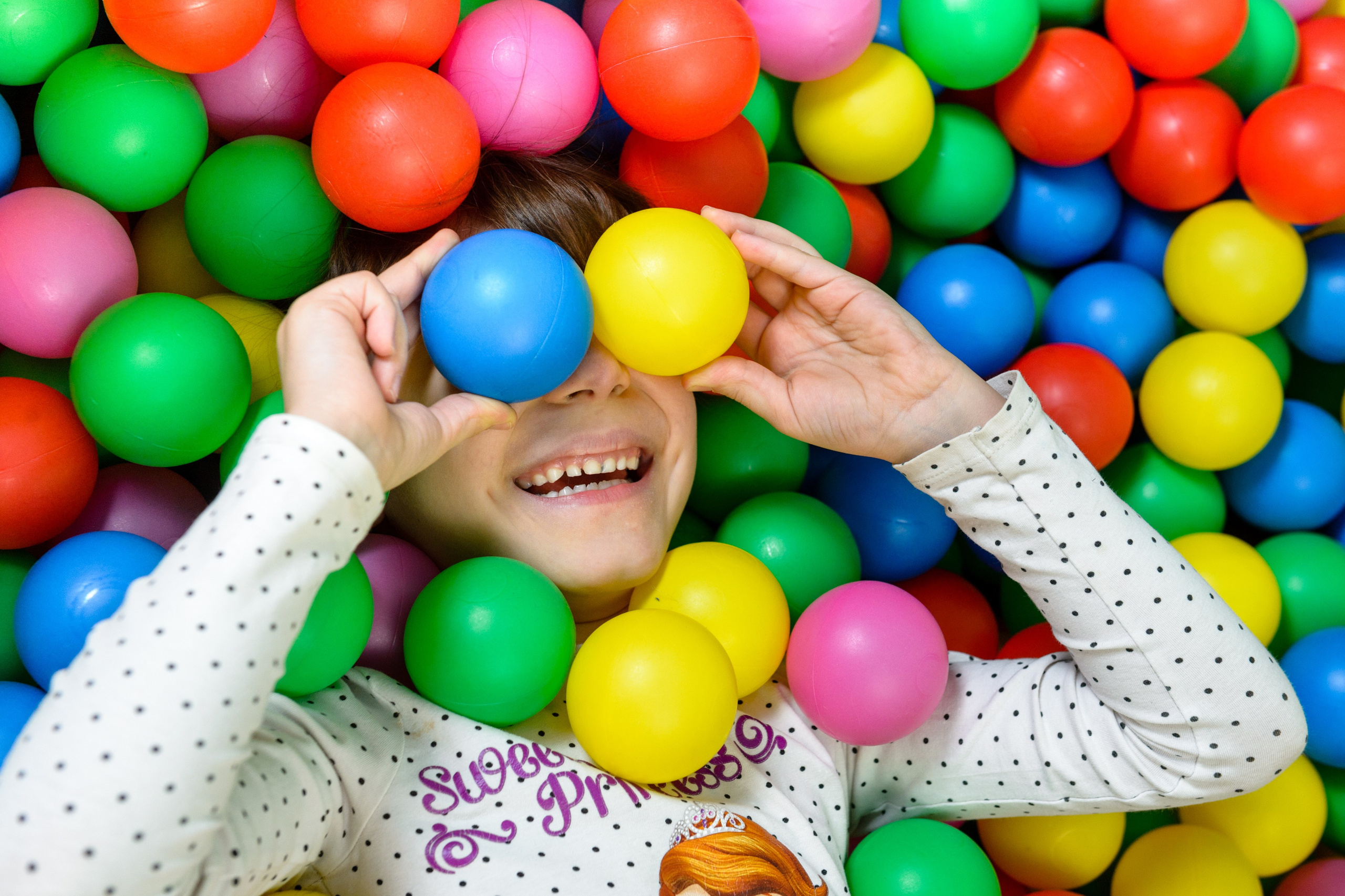 Вид сверху: ребёнок лежит в сухом бассейне среди разноцветных шариков.