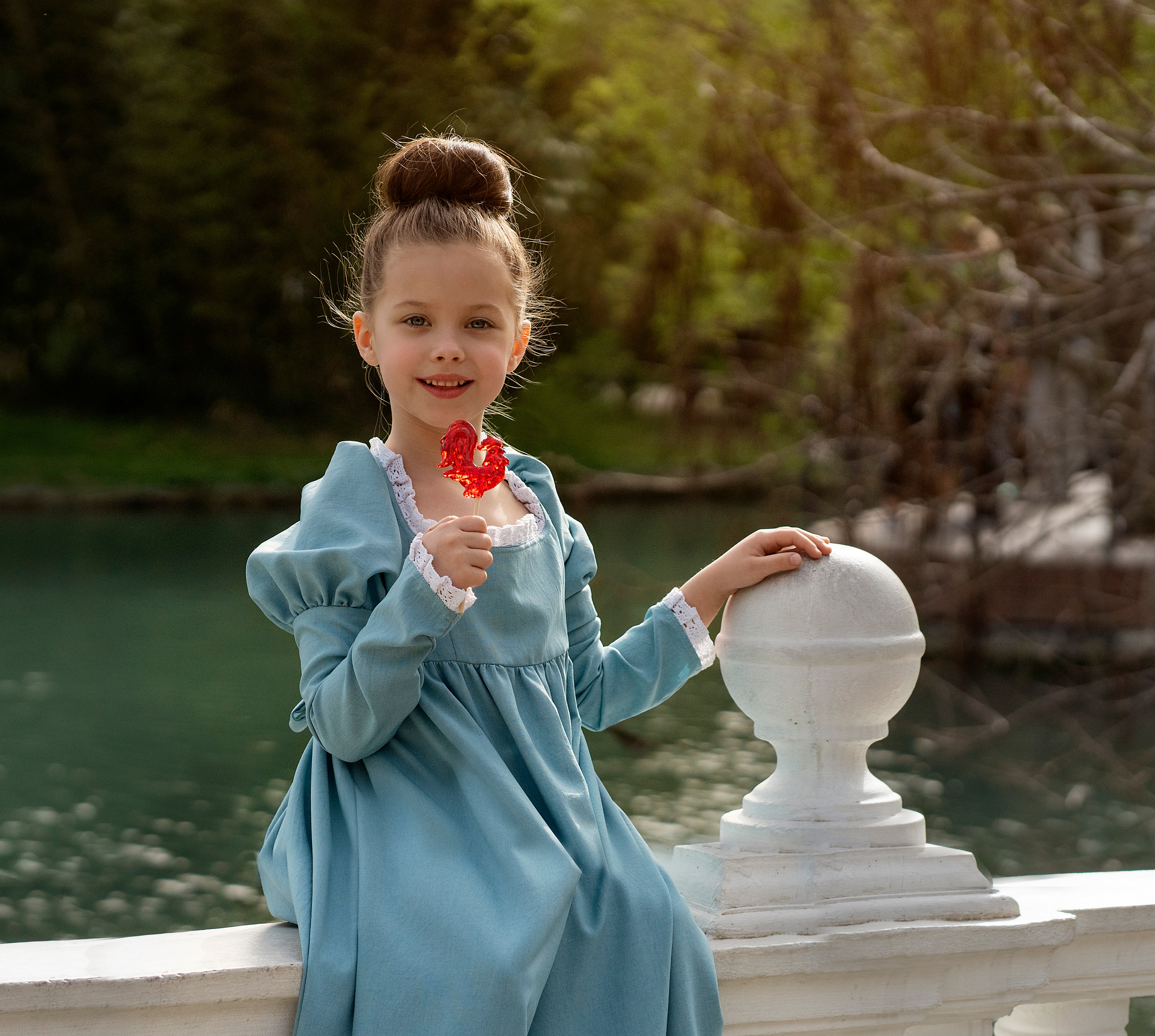 Девочка в старинном, голубом платье сидит на балясине и облизывает сладкого петушка на палочке.