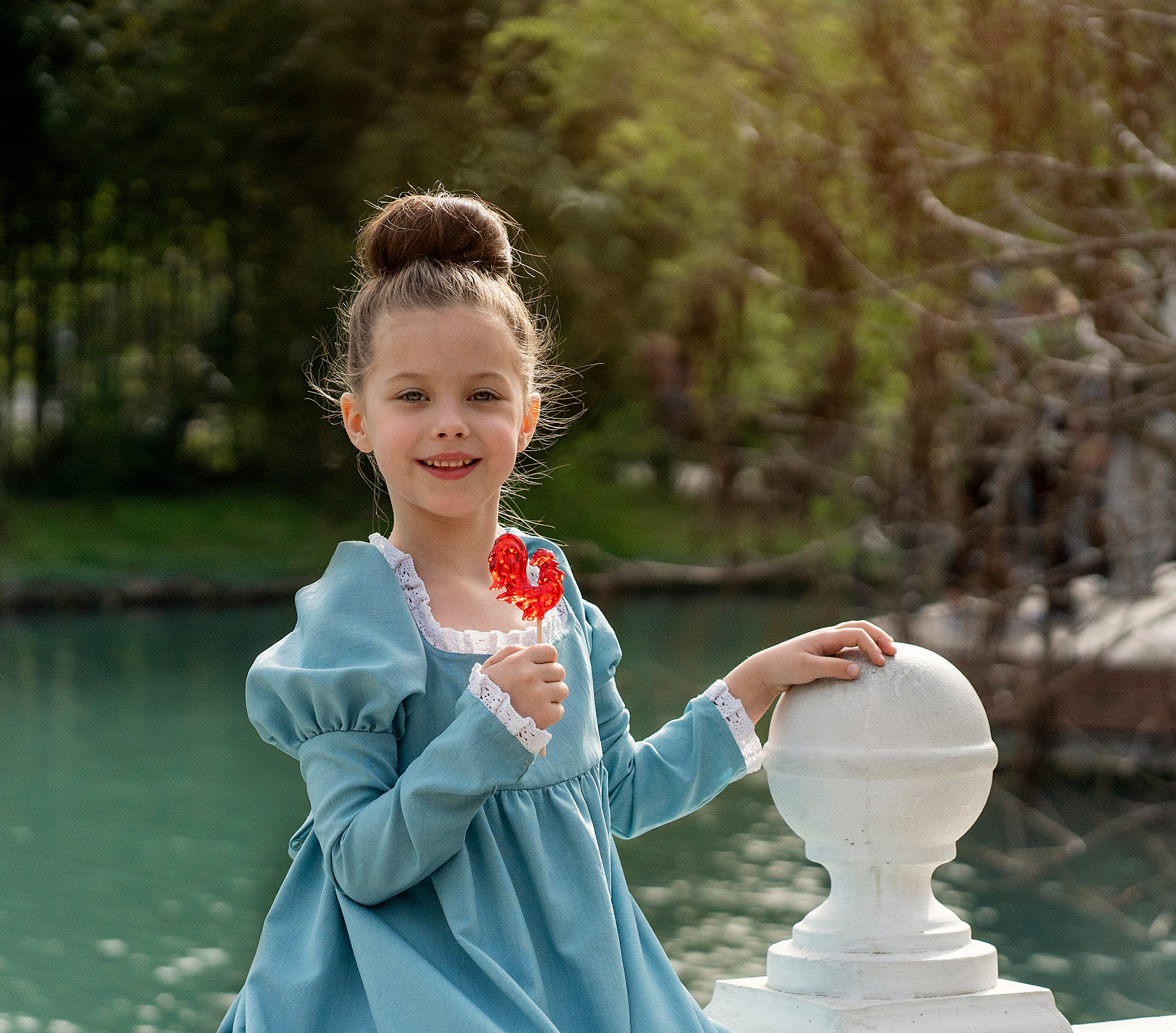 Девочка в голубом платье, в руках петушок на палочке. На заднем плане пруд.