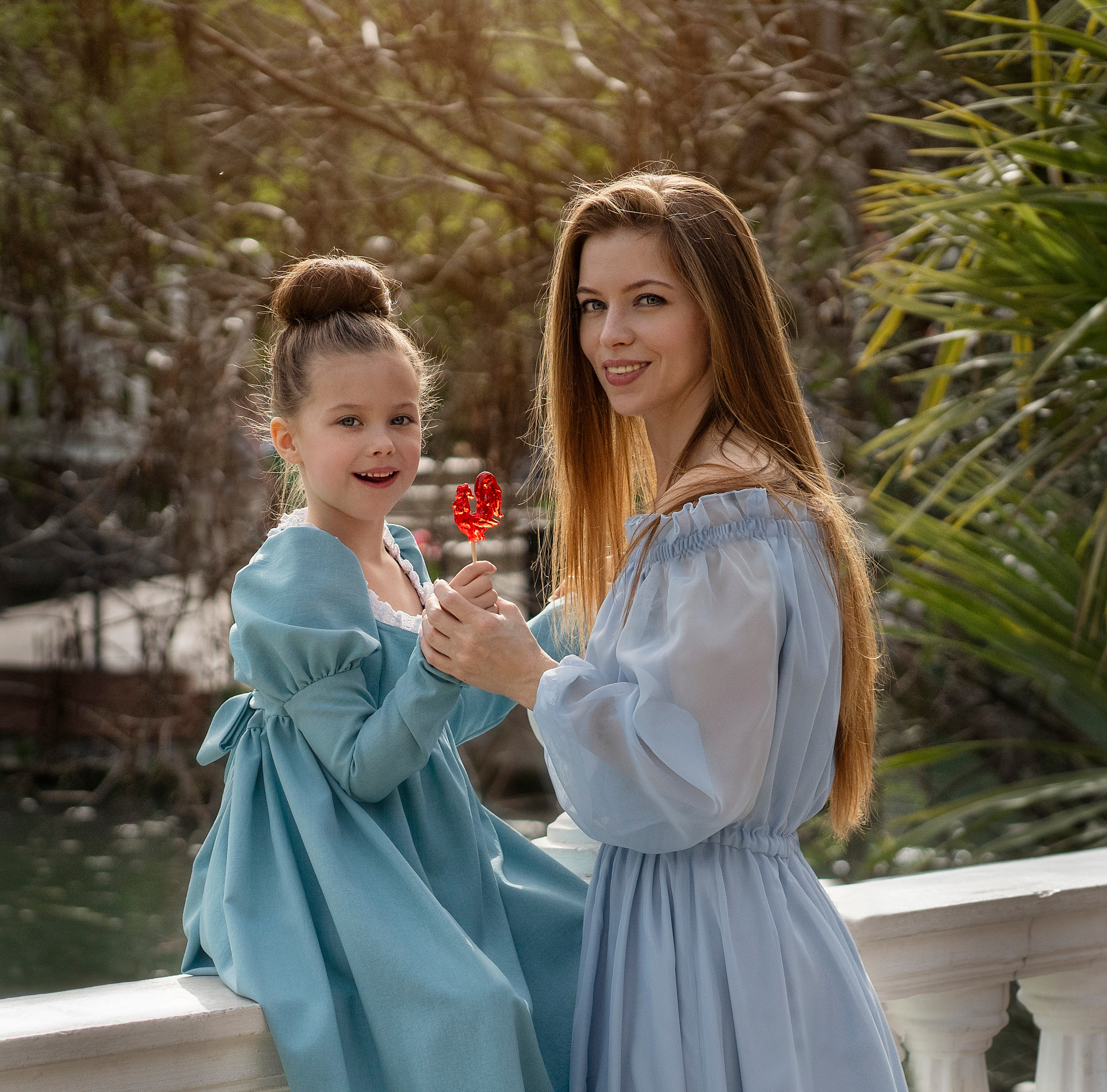 Мама угощает свою дочь петушком на палочке.