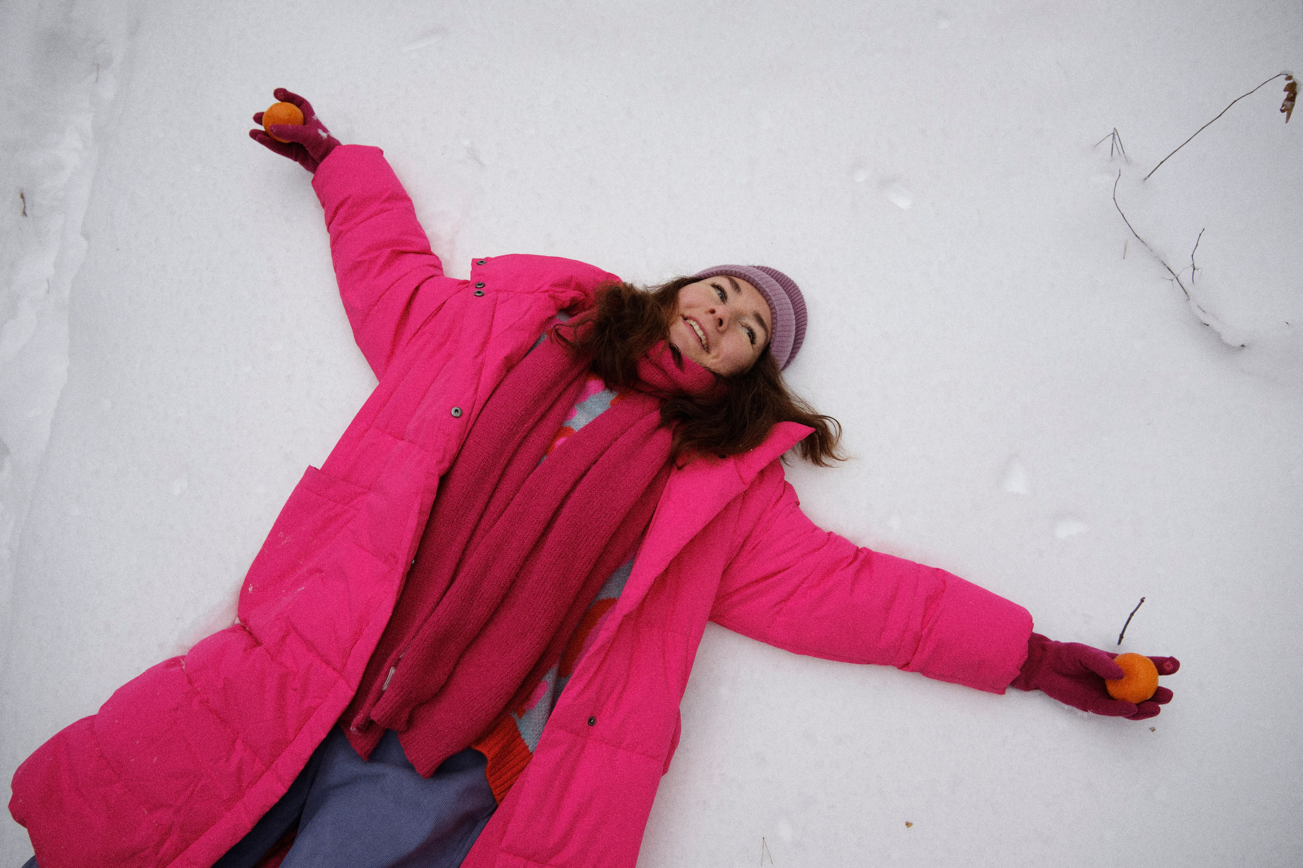 Женщина в яркой одежде лежит на снегу, раскинув руки с мандаринами и улыбается