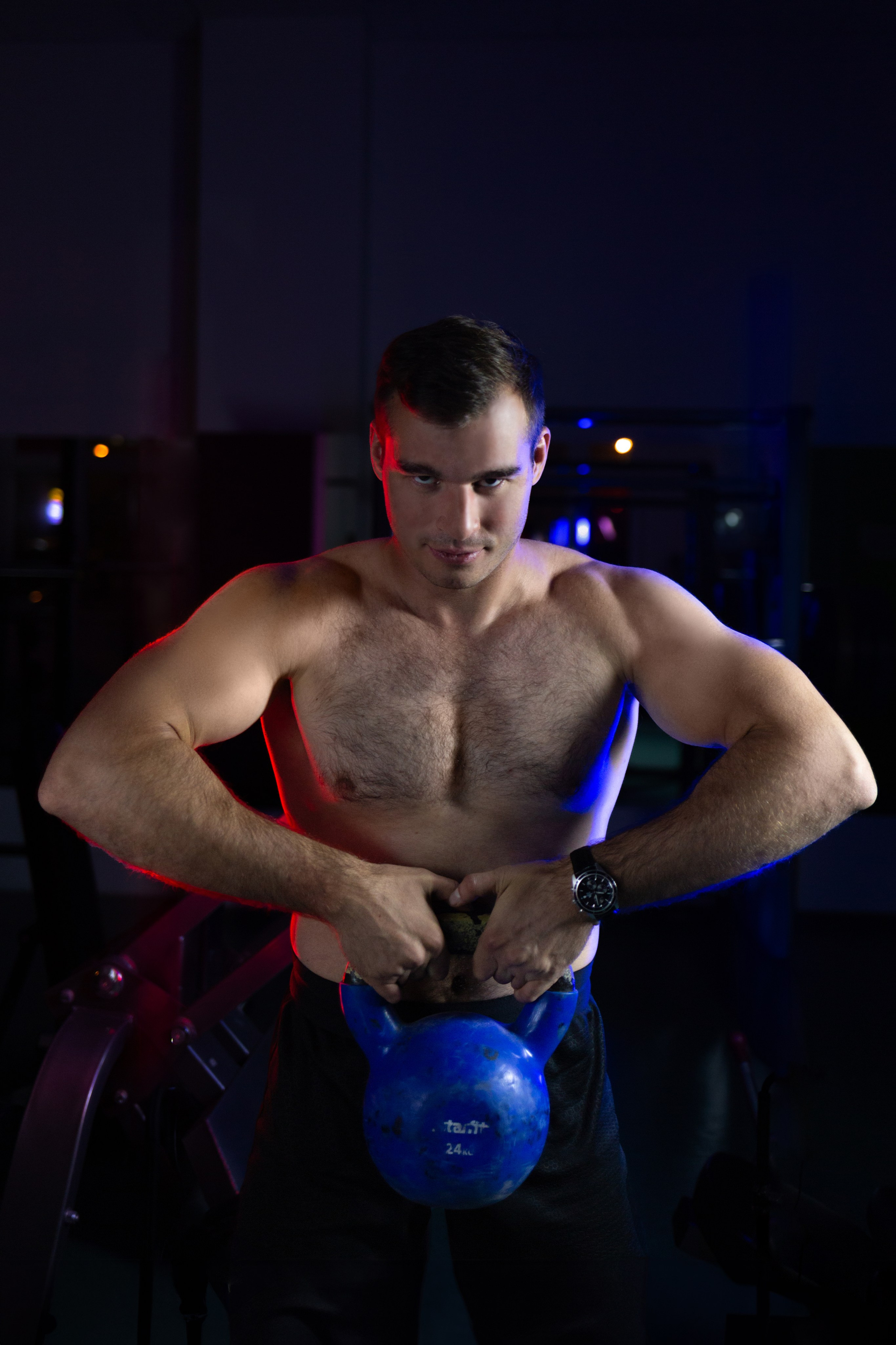 Голый по пояс мужчина держит гирю. Парень с красивыми мускулами в темных тонах с подсветкой.