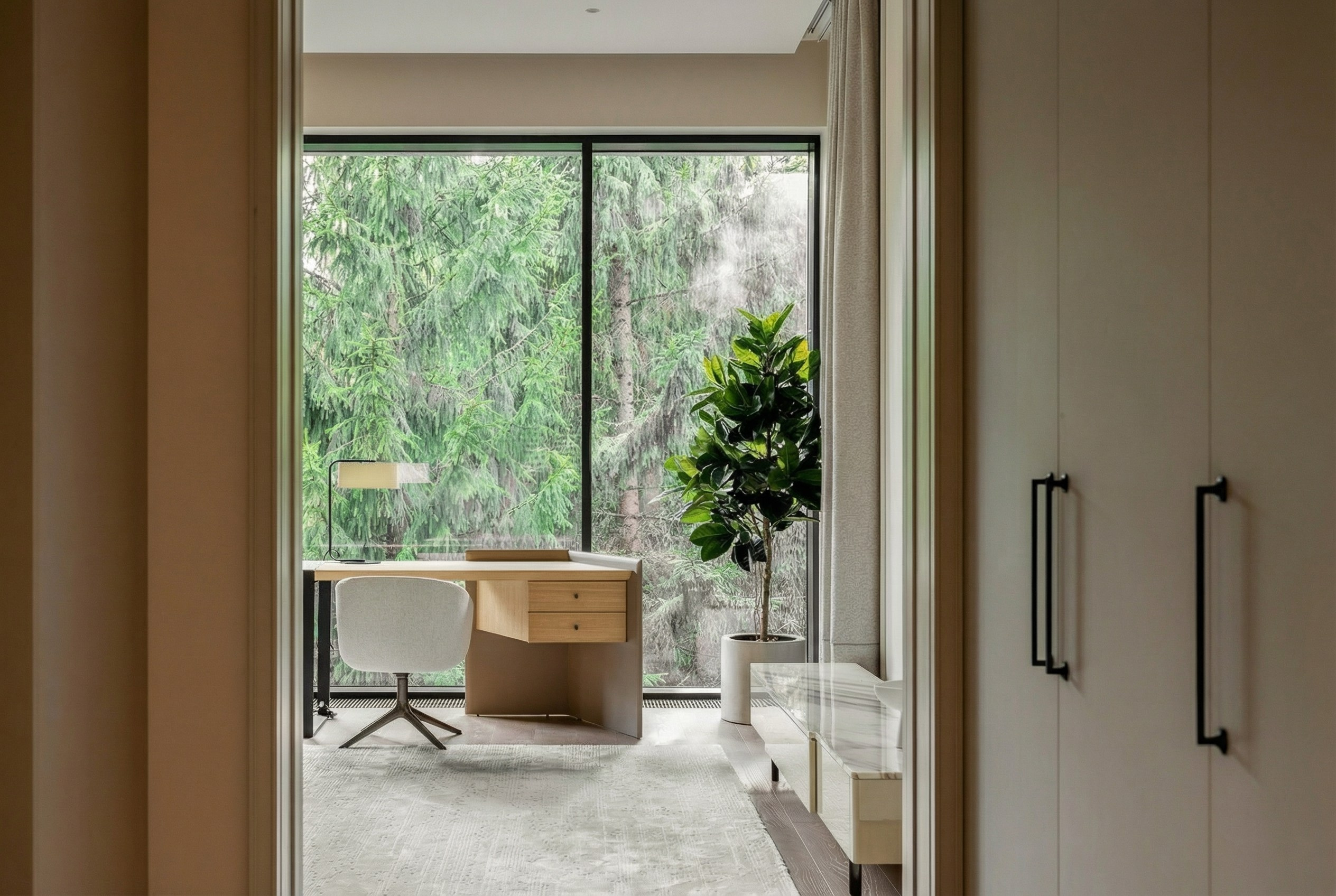 Вид на рабочее место в мастер-спальне через дверной портал, панорамное окно с видом на хвойный лес и встроенные системы хранения, проект Валерии Котович в Подмосковье.
