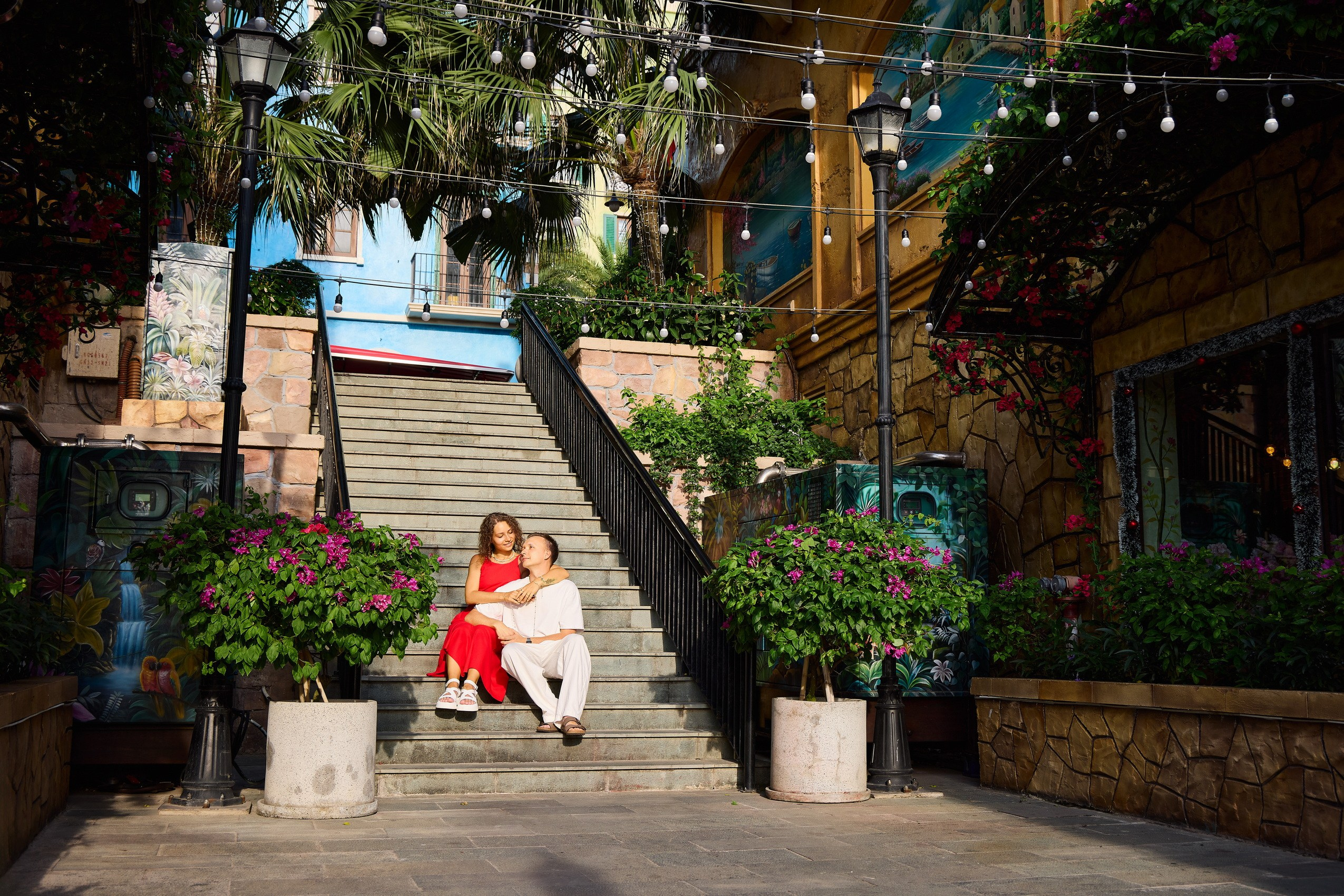 Couple in red and white clothes sitting on the stone Dragon Staircase surrounded by plants and Mediterranean architecture in Sunset Town, Phu Quoc, Vietnam.