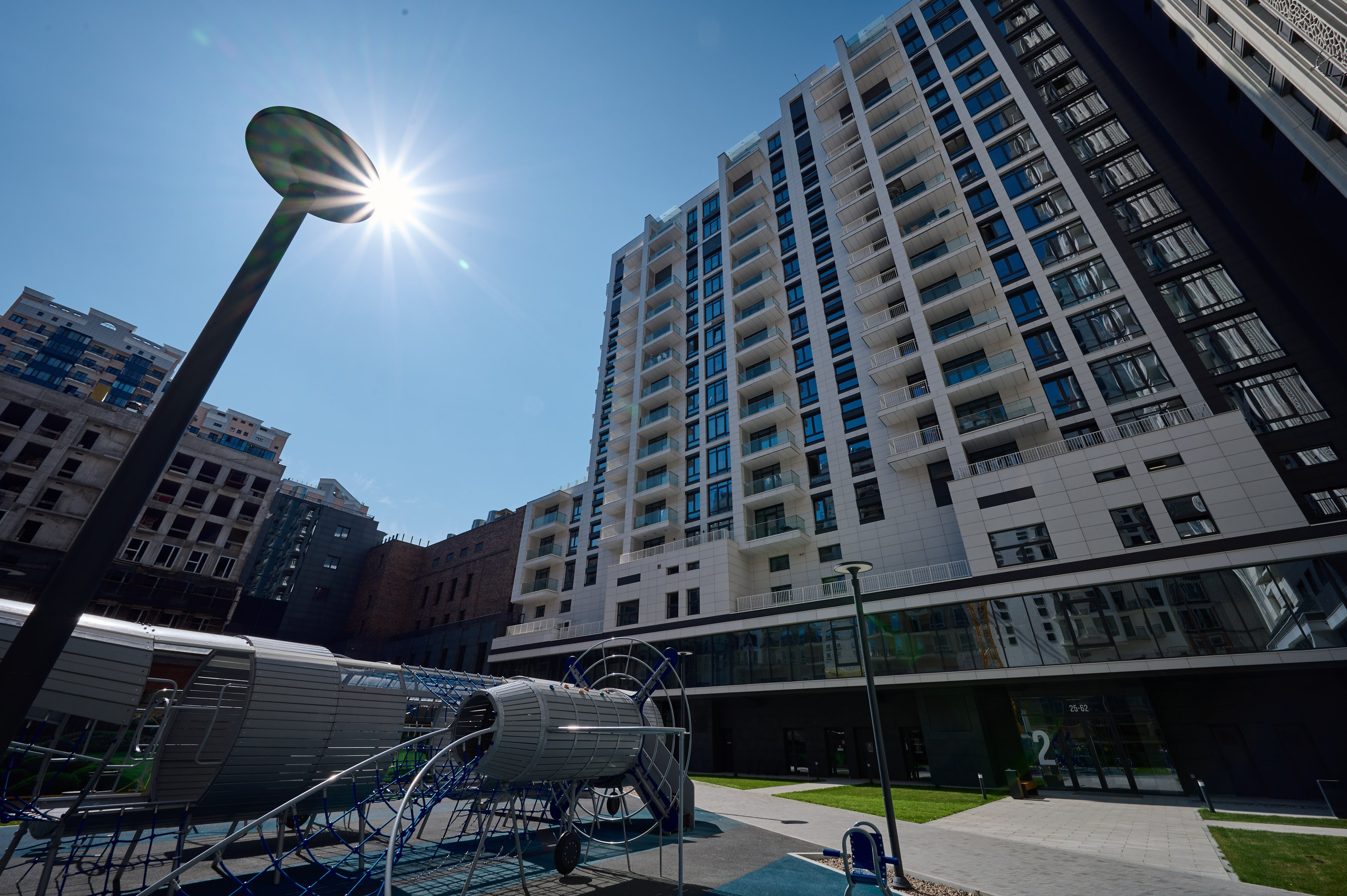 Вид на жилой комплекс с детской площадкой и уличным фонарём на переднем плане; солнце светит сквозь голубое небо, подчёркивая высоту зданий