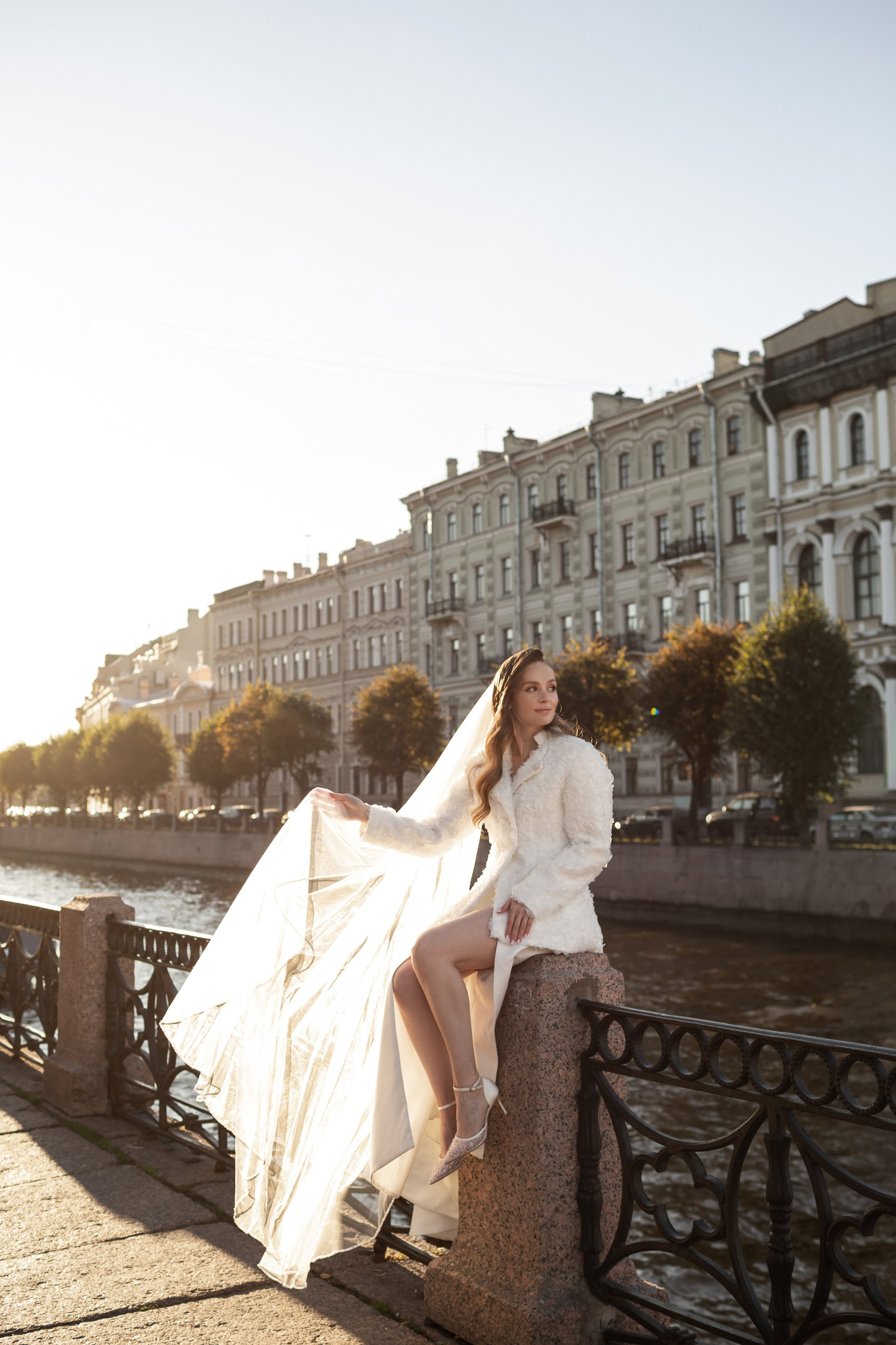 Невеста с длинной фатой и белым полушубком сидит на набережной.