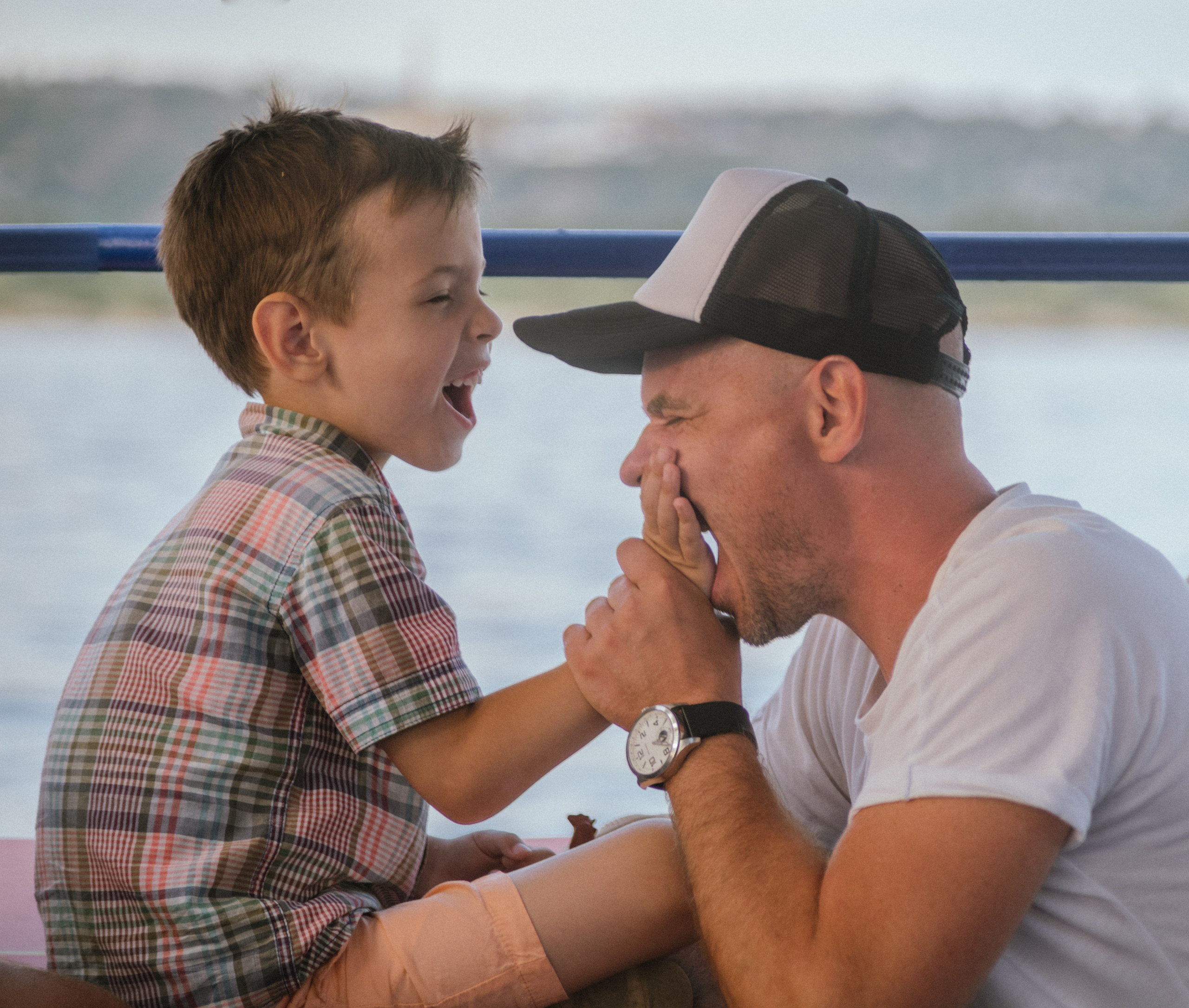 Сын дразнит отца, оба смеются и играют, сидя рядом во время речной прогулки — искренний момент близости.