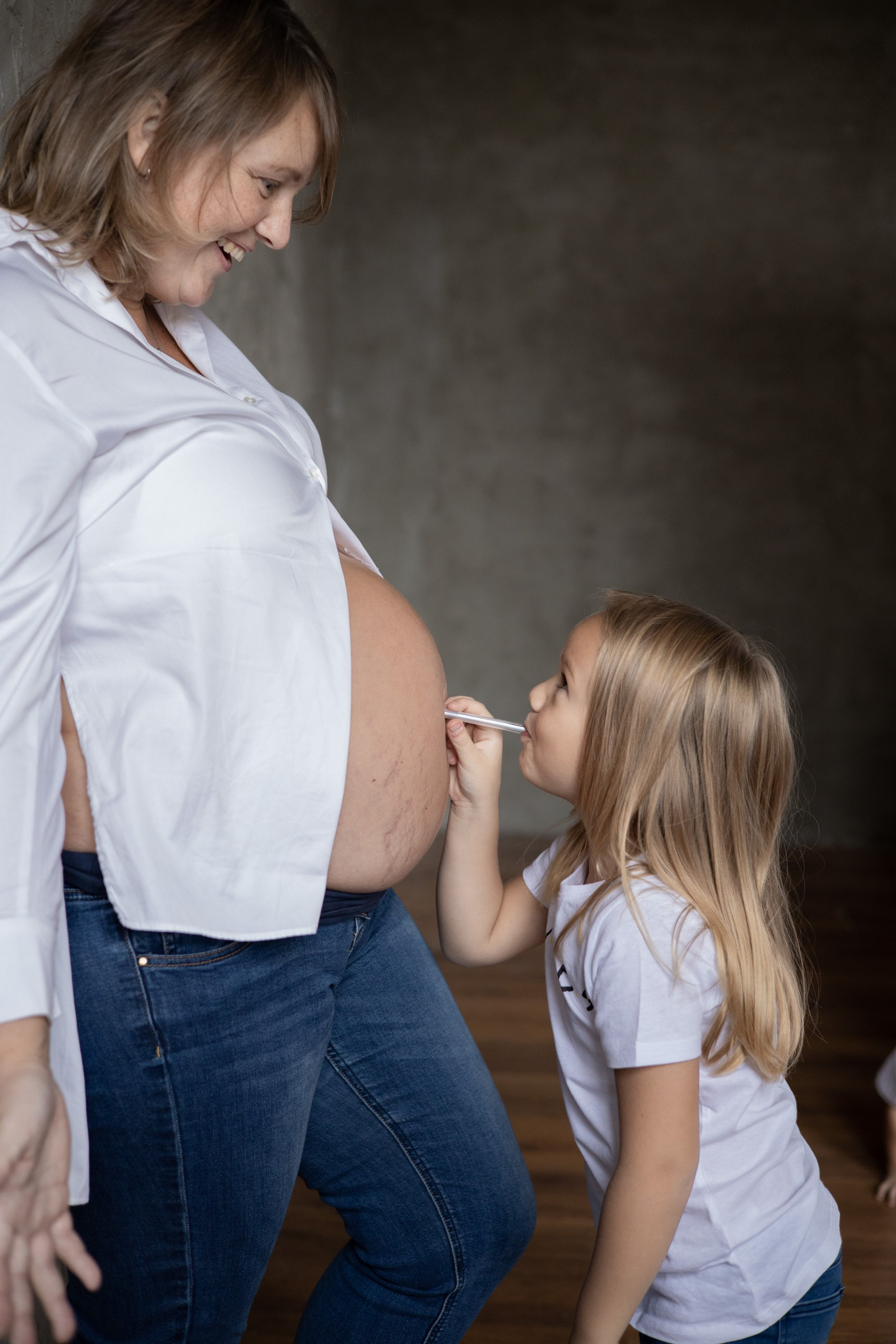Маленькая девочка "надувает" мамин живот через соломинку. Беременная мама стоит у стены. Фотосессия для беременных  Астрахань.