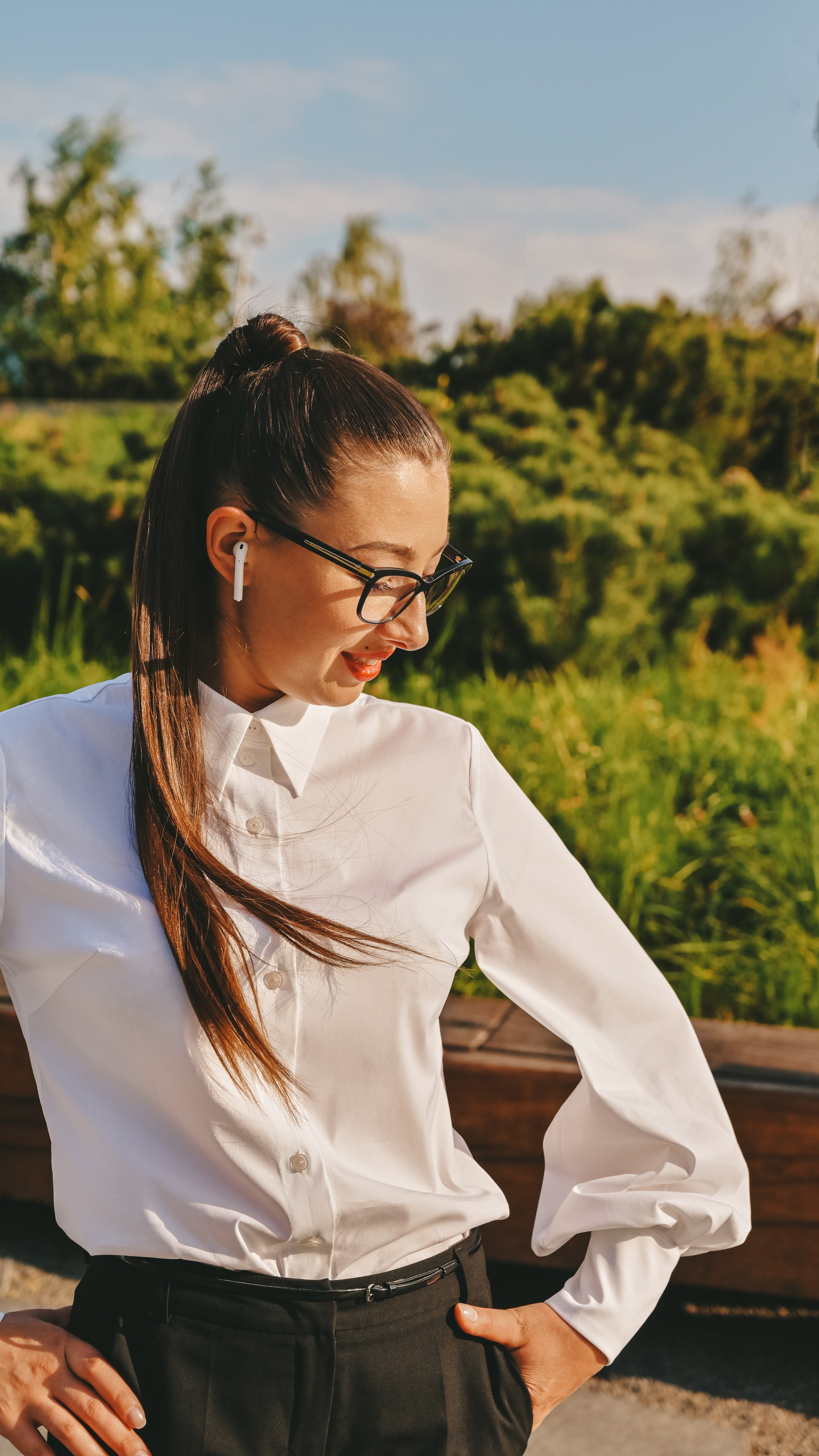 Молодая женщина в деловом и элегантном образе с белой блузкой, черными брюками и черными очками стоит на фоне живописного паркового пейзажа. Ее длинные волосы аккуратно уложены в высокий хвост, что придает ей современный и профессиональный вид. Наушники в ухе добавляют элемент современности и стиля. Она одета в строгий комплект, который идеально сочетается с окружающей природной средой. Светлый фон неба и зеленые деревья создают ощущение свежести и легкости. Этот снимок демонстрирует мастерство работы с освещением и композицией, делая акцент на игре света и тени, а также на гармоничном сочетании человека и природы. Perfect for business and lifestyle photography enthusiasts!