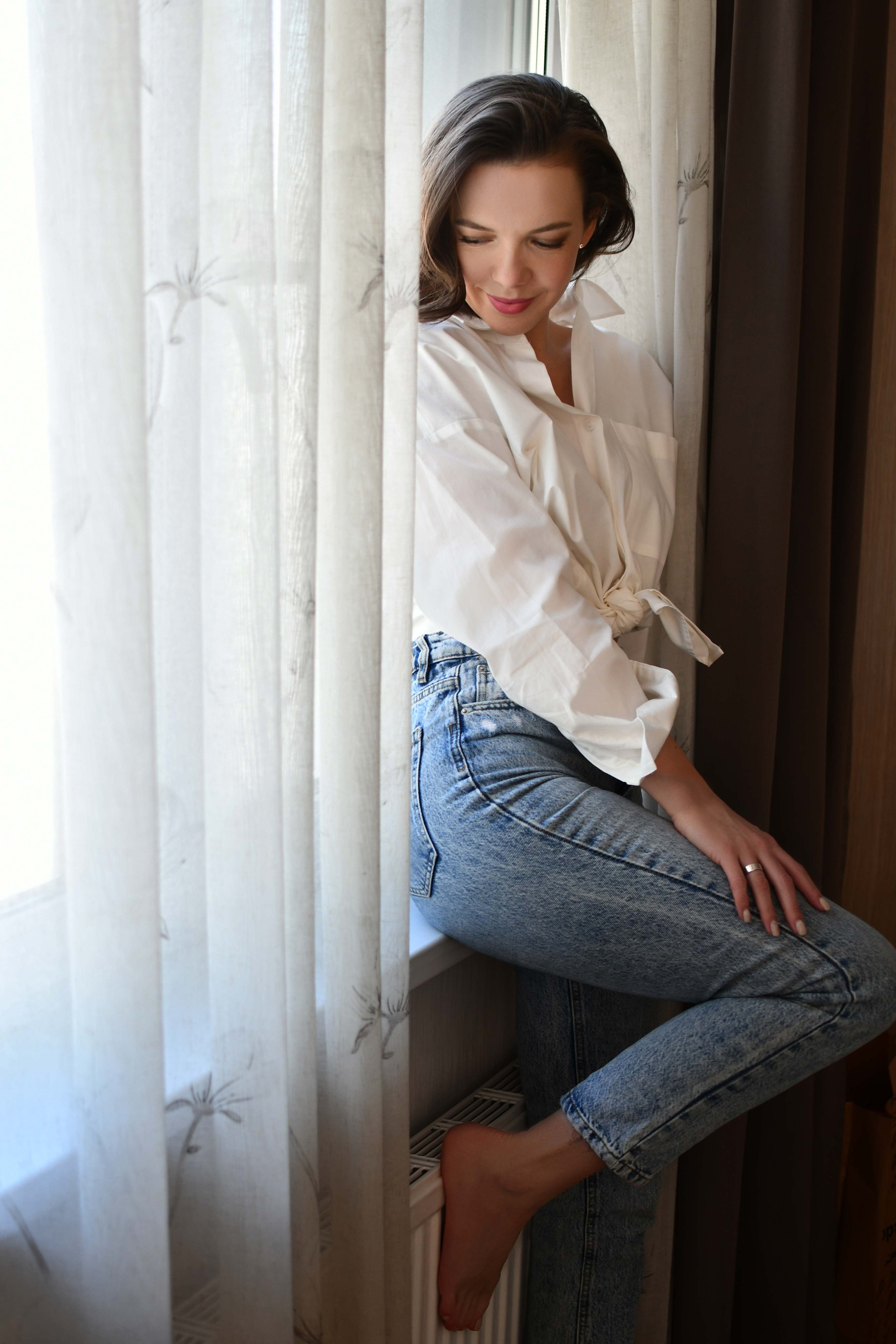 Девушка в белой рубашке и голубых джинсах стоит у окна