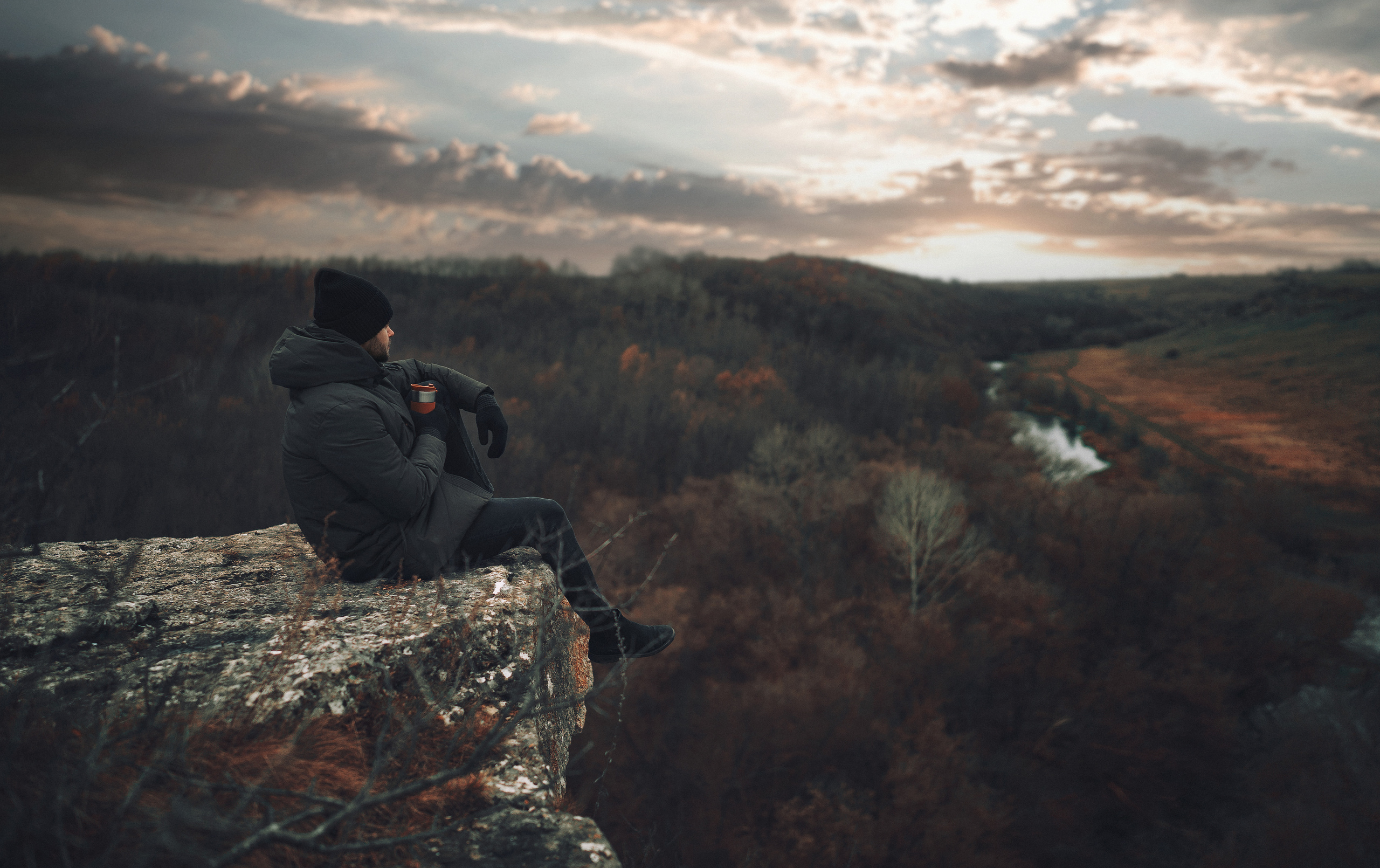 Парень в шапке и теплой куртке сидит на краю скалы.
