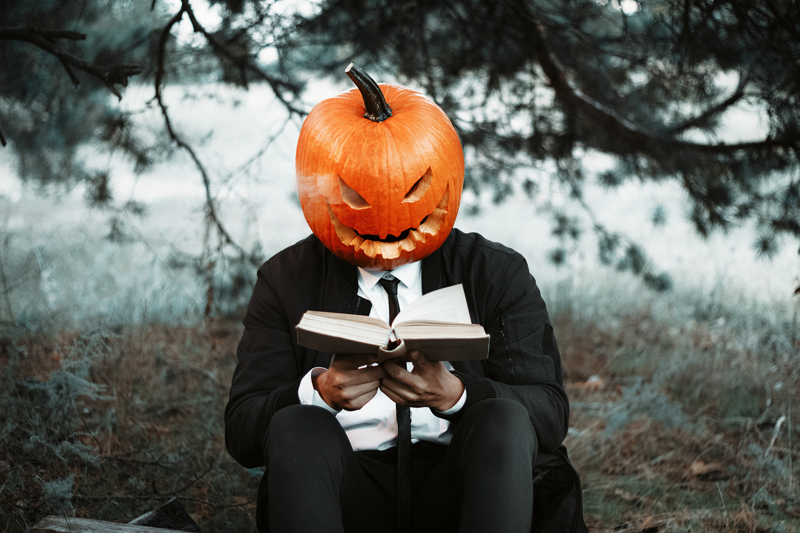 Парень в строгом костюме в маске тыквы читает книгу и смотрит на нас