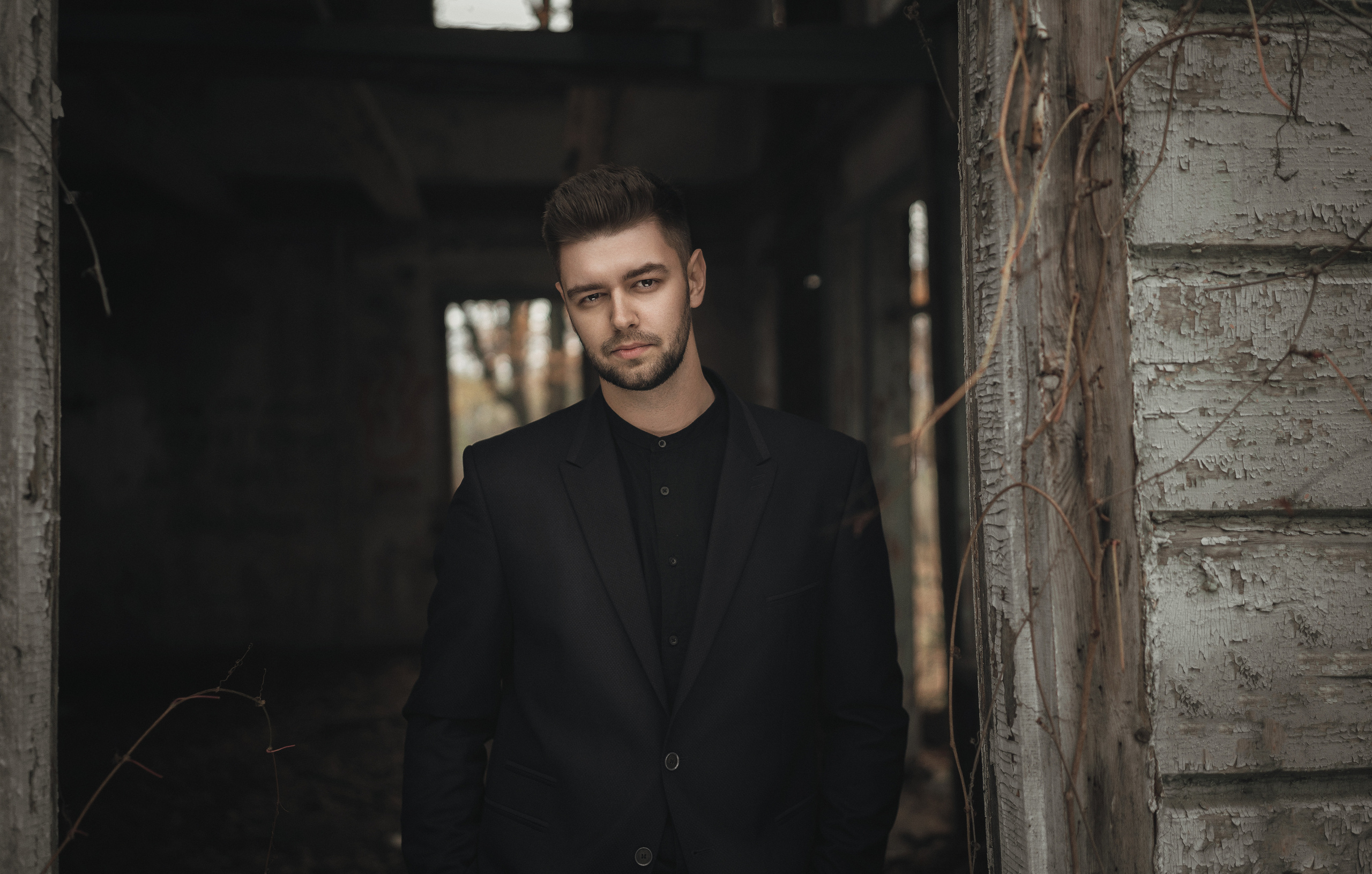 Парень в черном строгом костюме стоит на улице между створок окон.