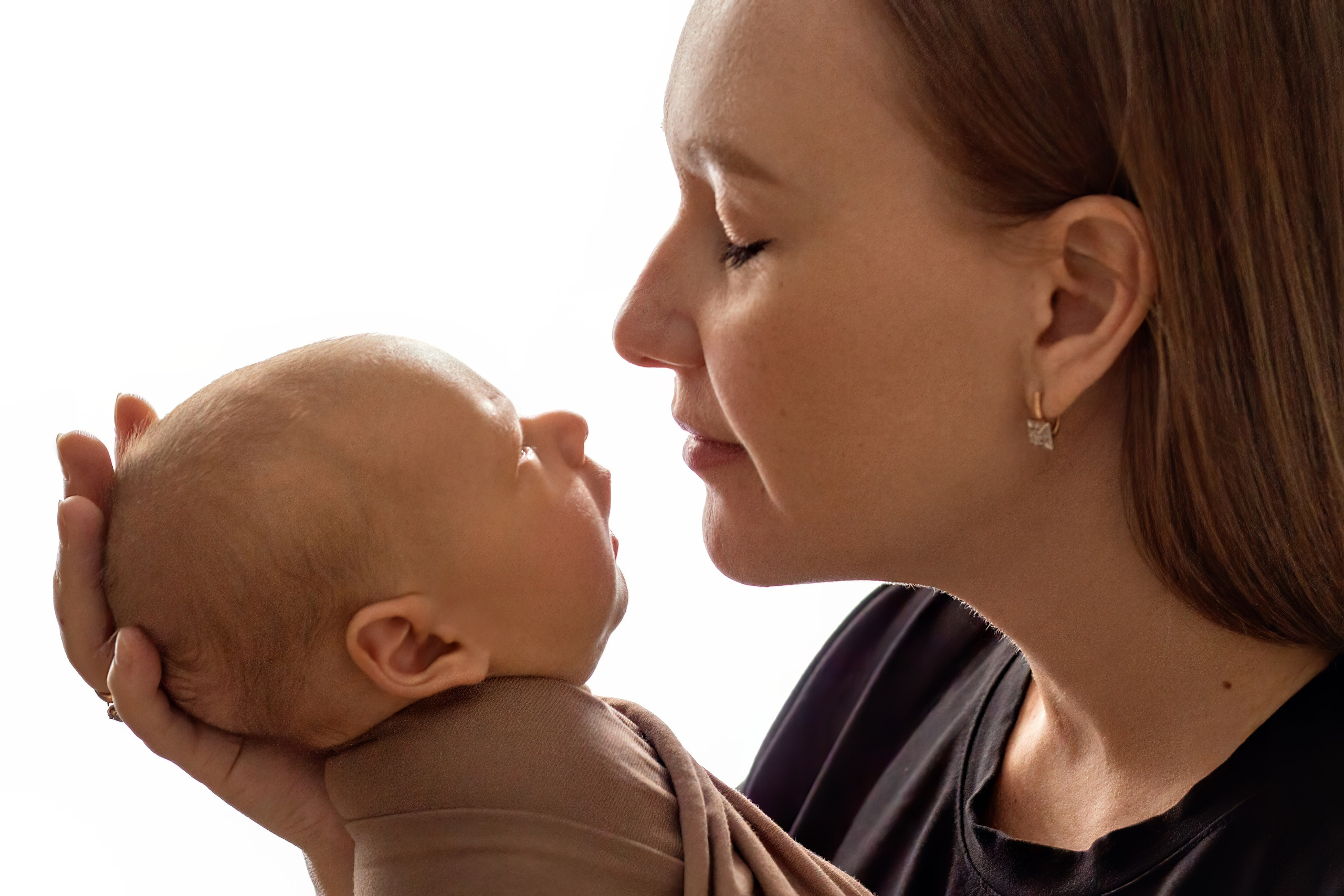 крупный портрет молодой мамы в черной футболке нюхающей своего малыша укутанного в бежевую ткань на белом фоне