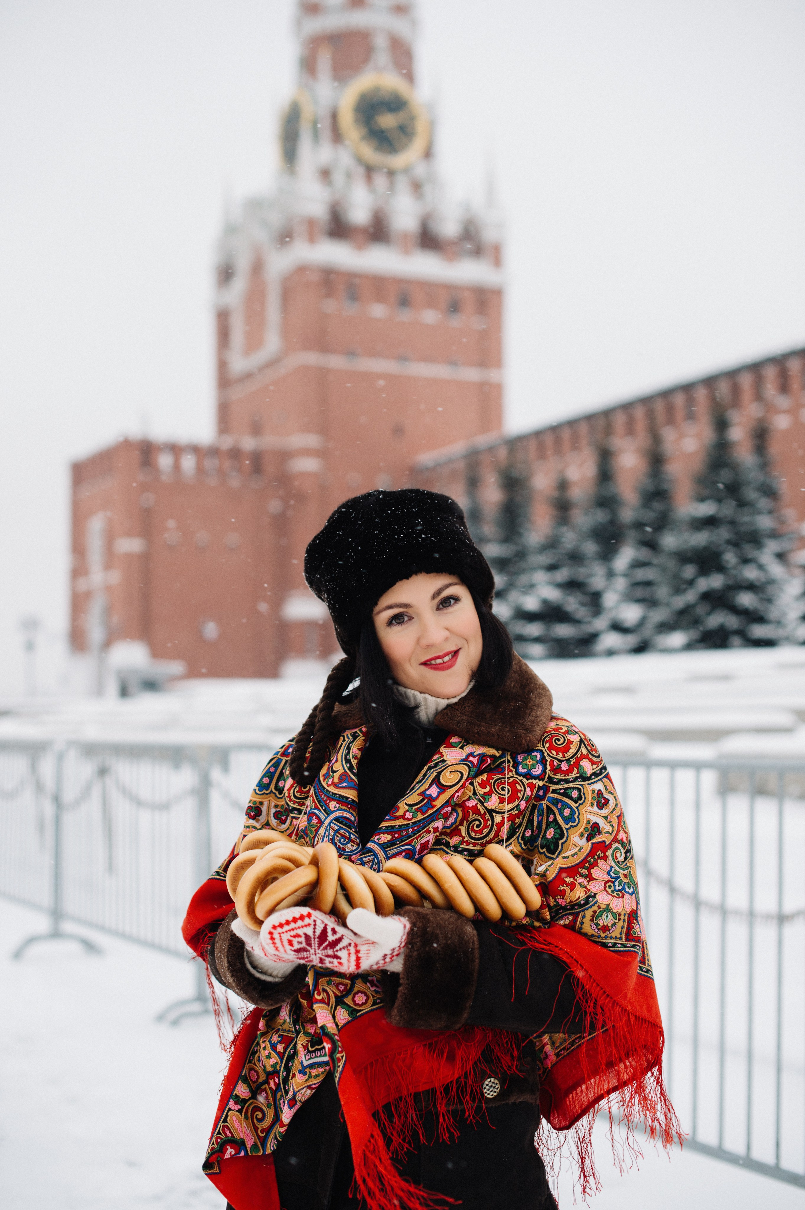 Девушка в красивом наряде в русском стиле с красным платком на плечах на фоне Спасской башни Кремля на Красной площади зимой. Она держит в руках вязанку баранок.