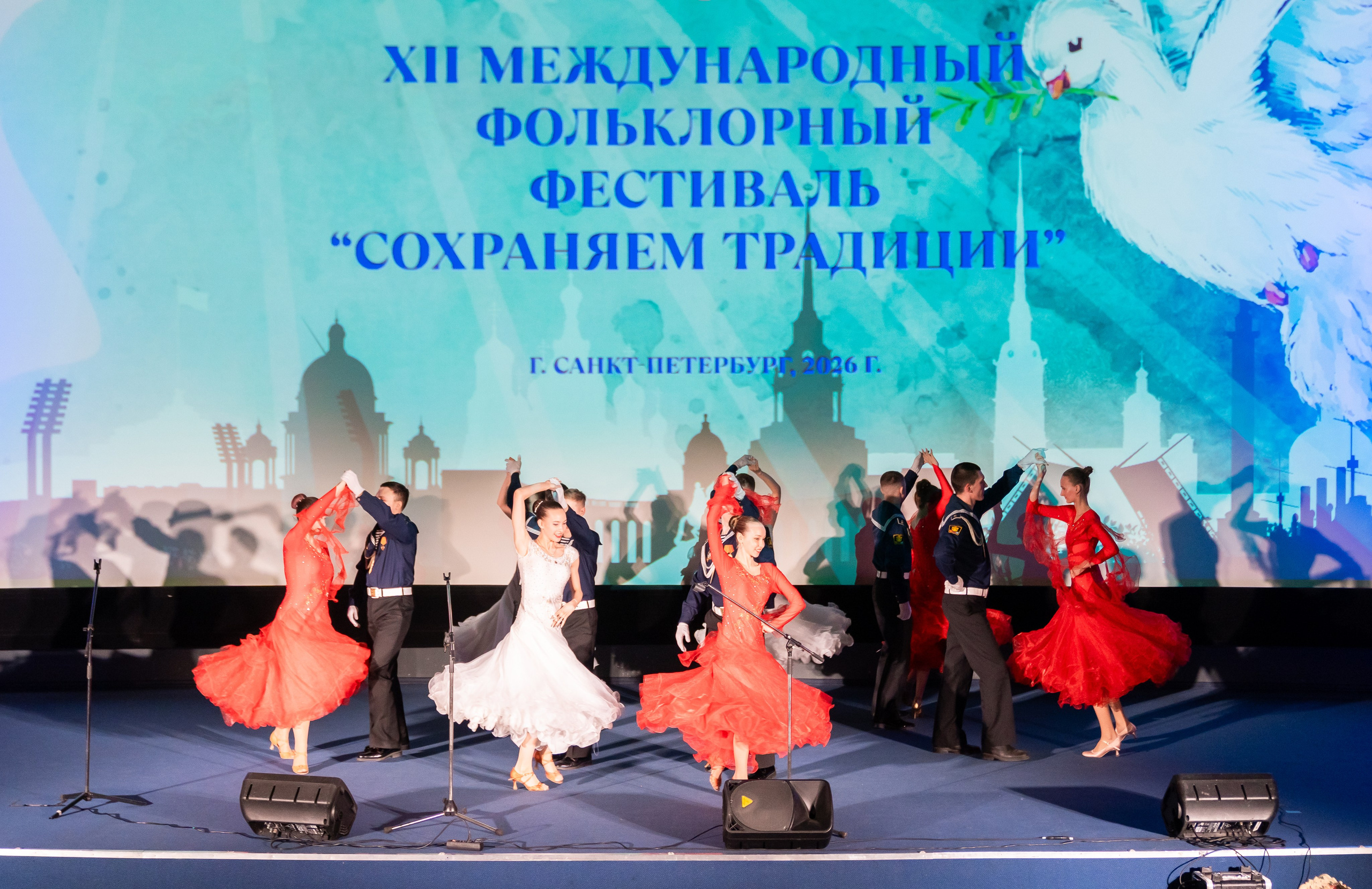 Танцевальный номер на сцене XII Международного фольклорного фестиваля «Сохраняем традиции» Санкт-Петербург 2026: девушки в красных и белых платьях, юноши в морской форме. Фото: Мальцев Евгений, evrednix.ru.